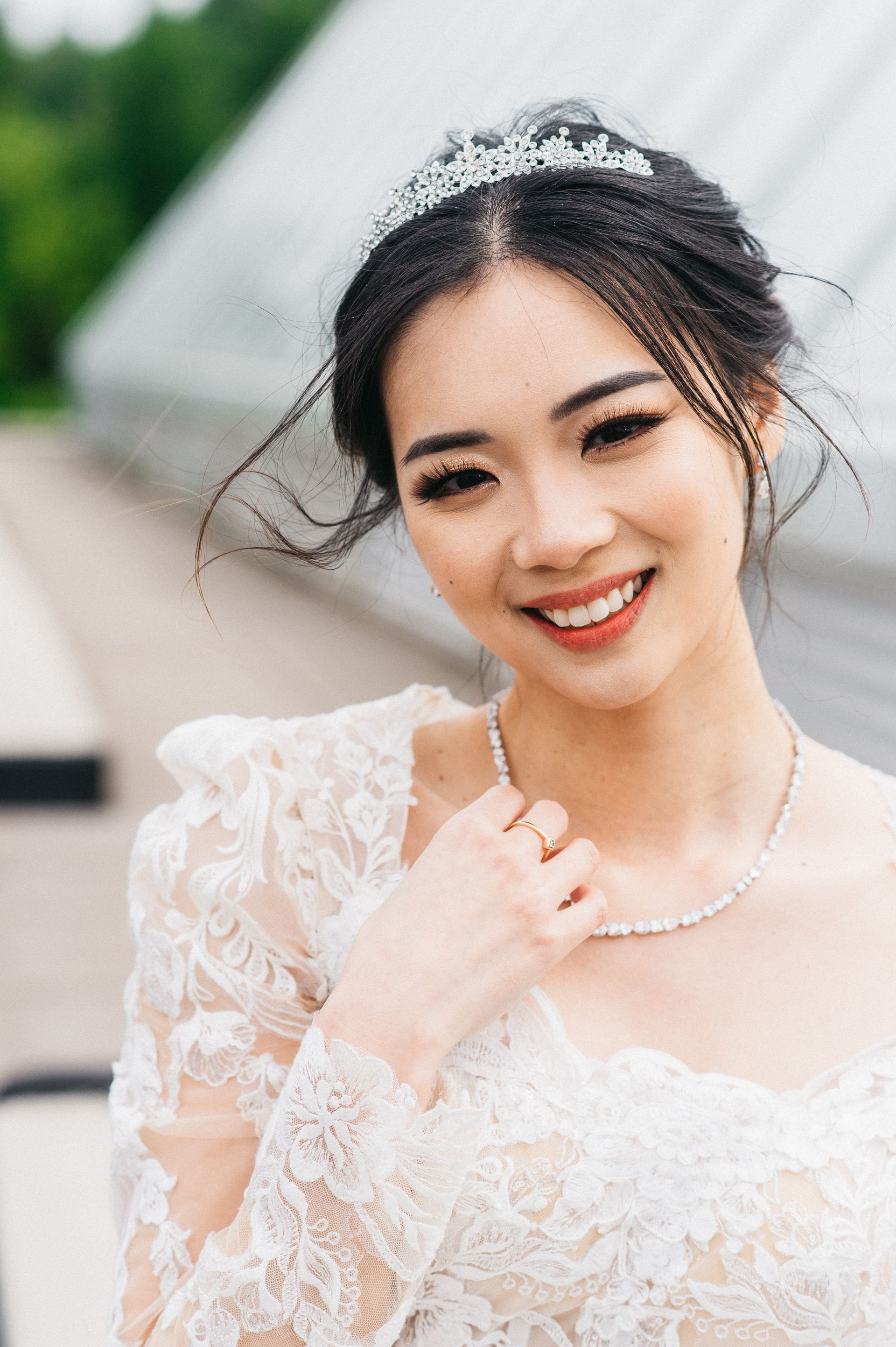 Woman in a white lace dress with a tiara and necklace smiling outdoors.