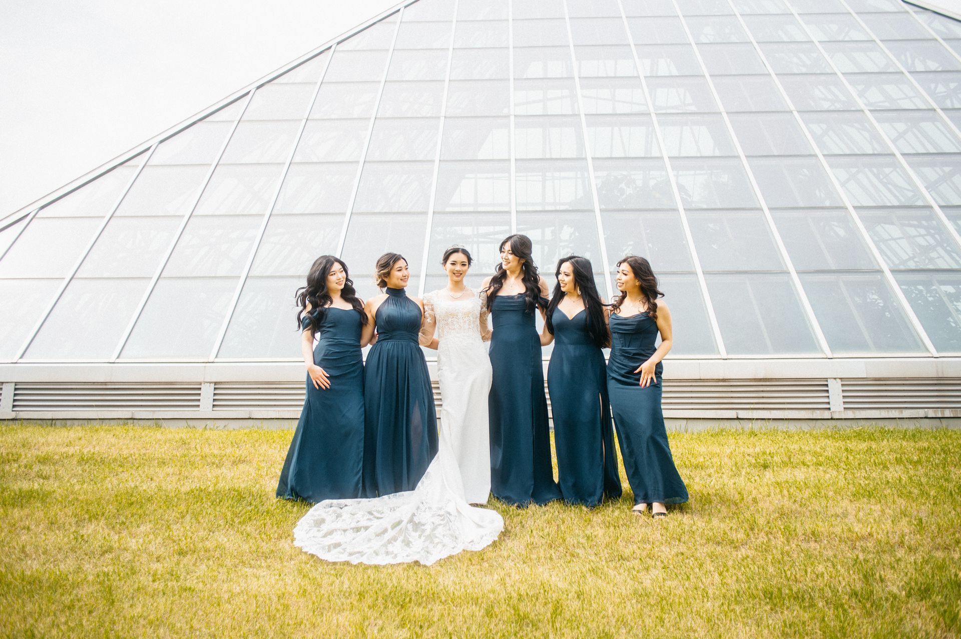 Bride and bridesmaids in navy gowns pose outdoors in front of a glass building.