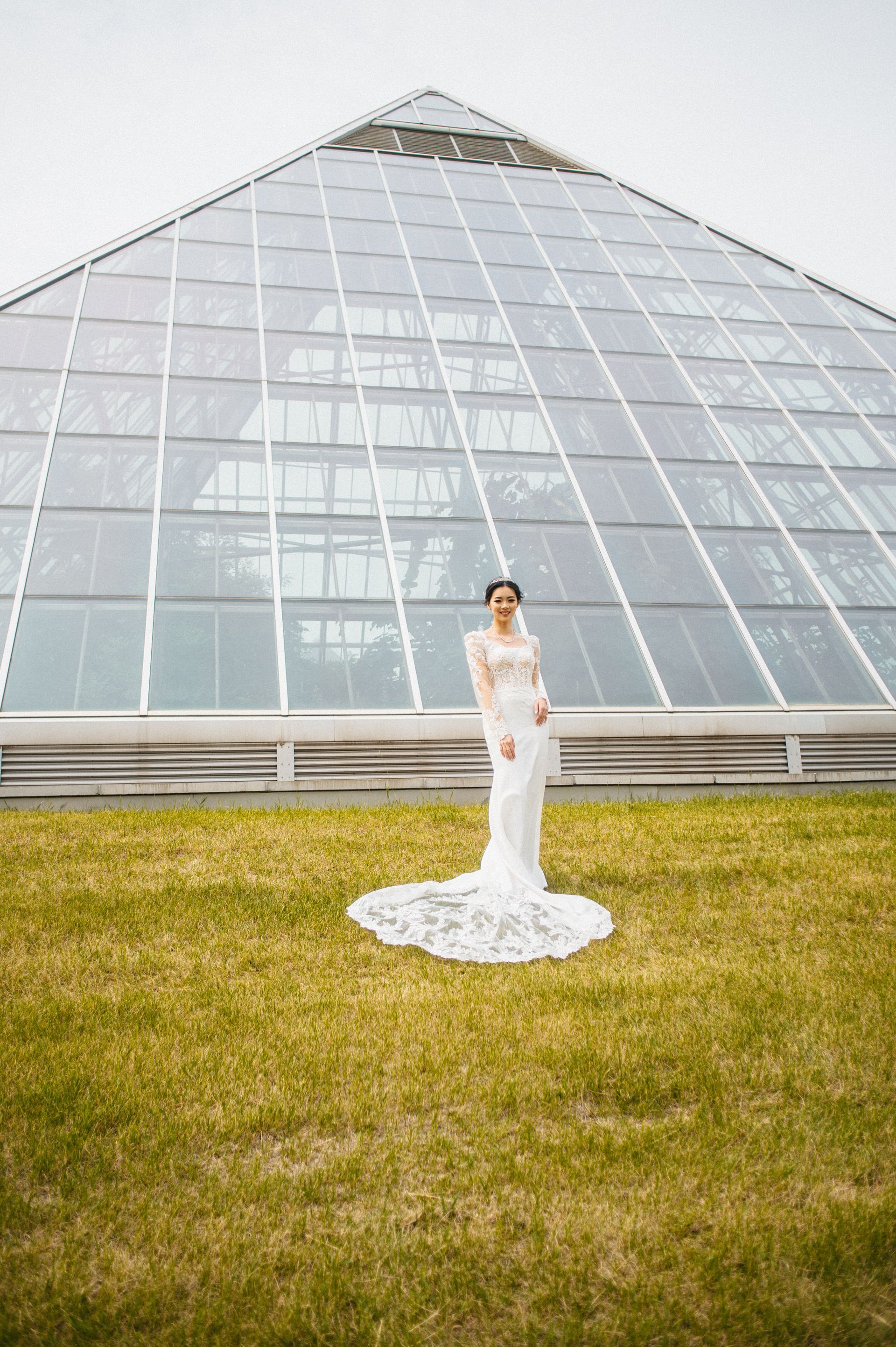Bride in a white lace wedding dress stands on grass in front of a glass pyramid greenhouse.