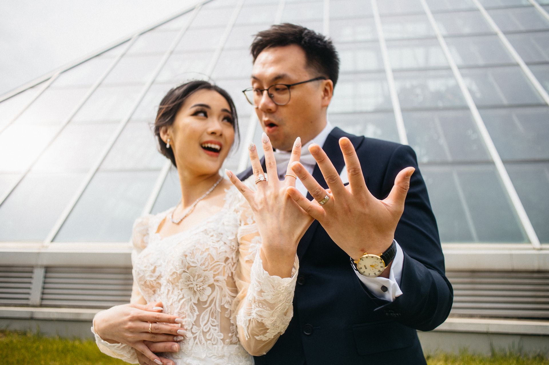Newlyweds show off wedding rings, surprised expressions. Outside, glass-paneled building in background.