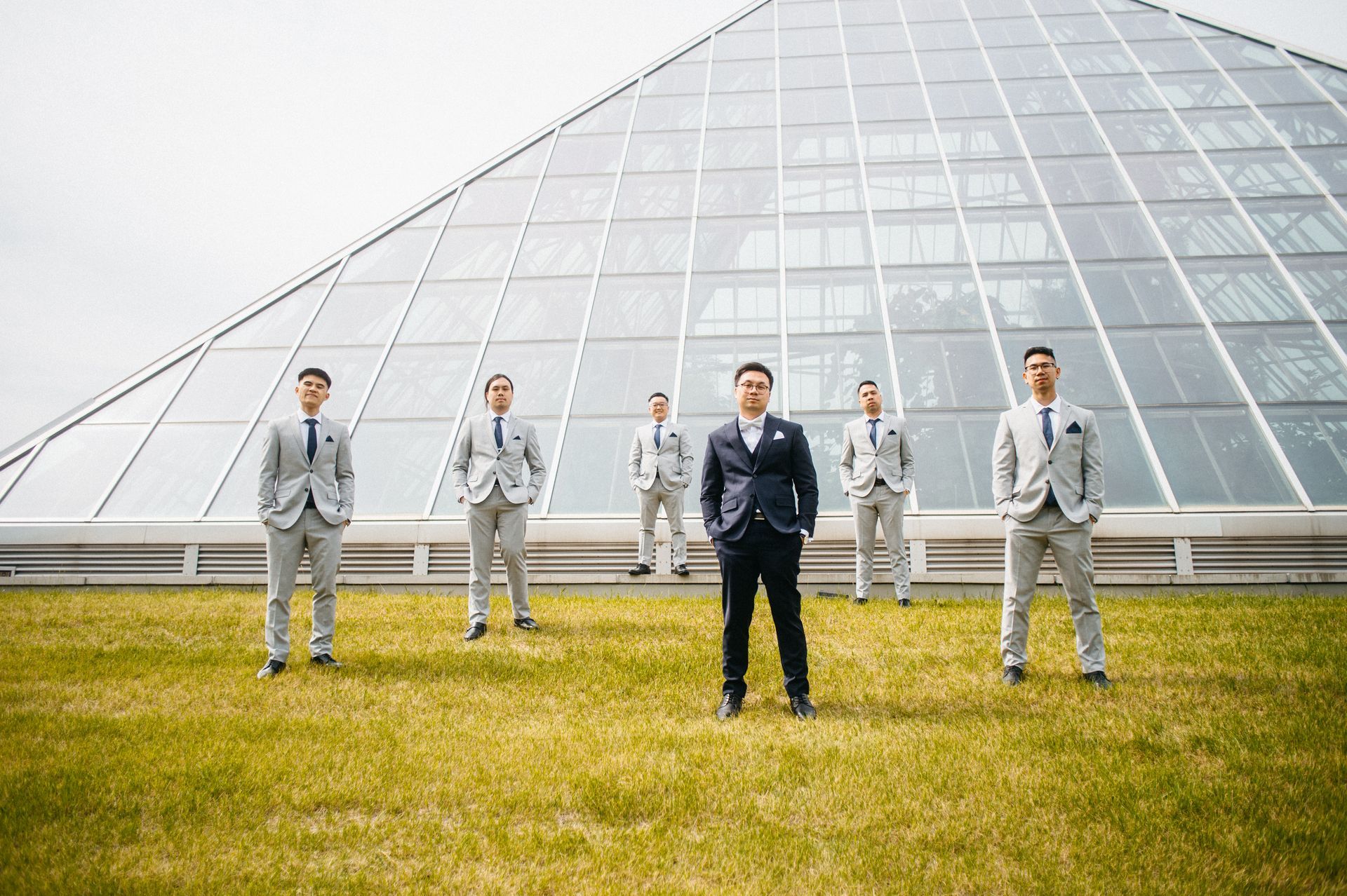 Six men in suits stand in front of a glass pyramid. One man in a navy suit, others in light gray.