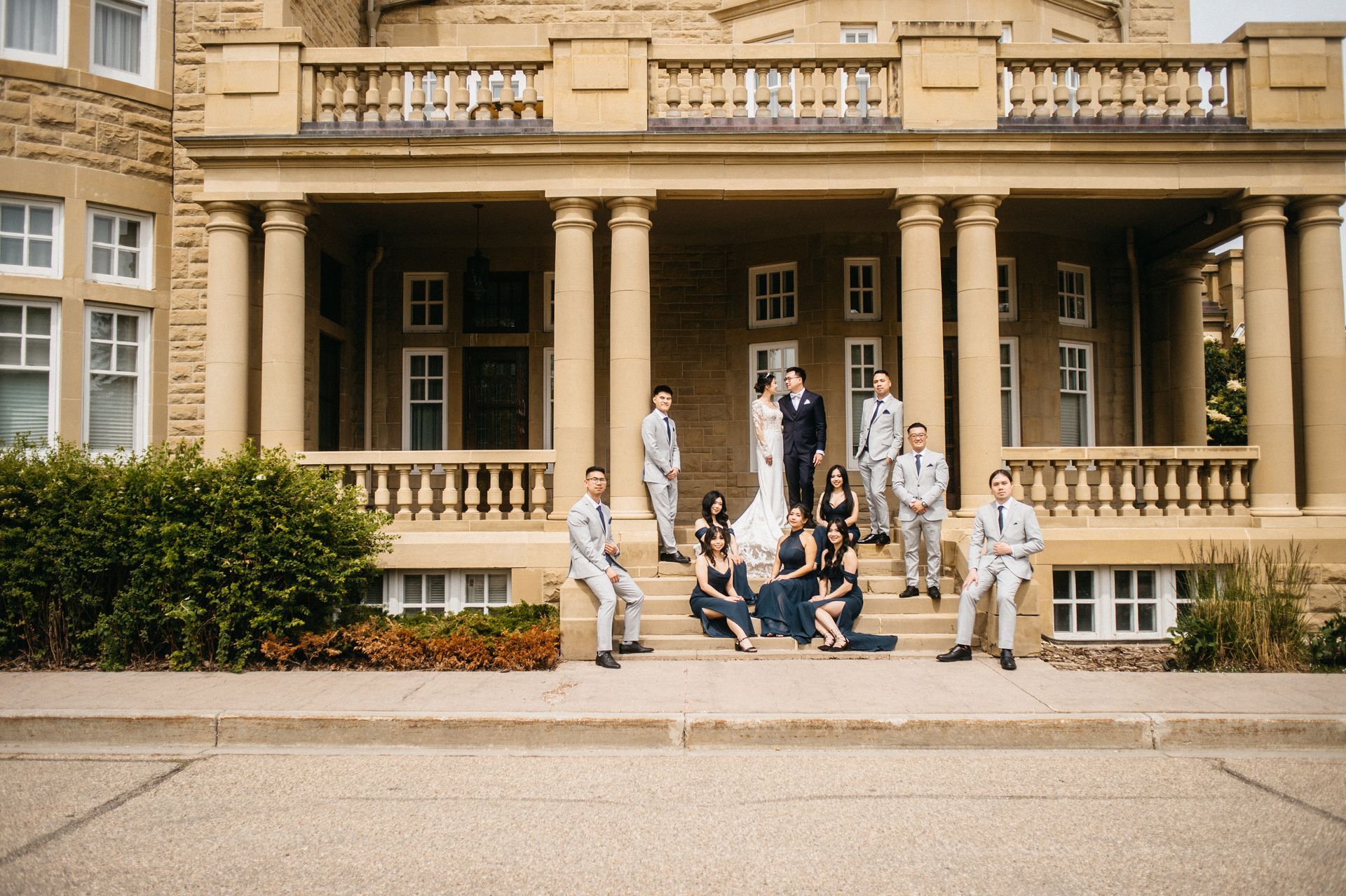 Wedding party poses on steps of a stone building with columns. Bride and groom in center, others in suits and dresses.