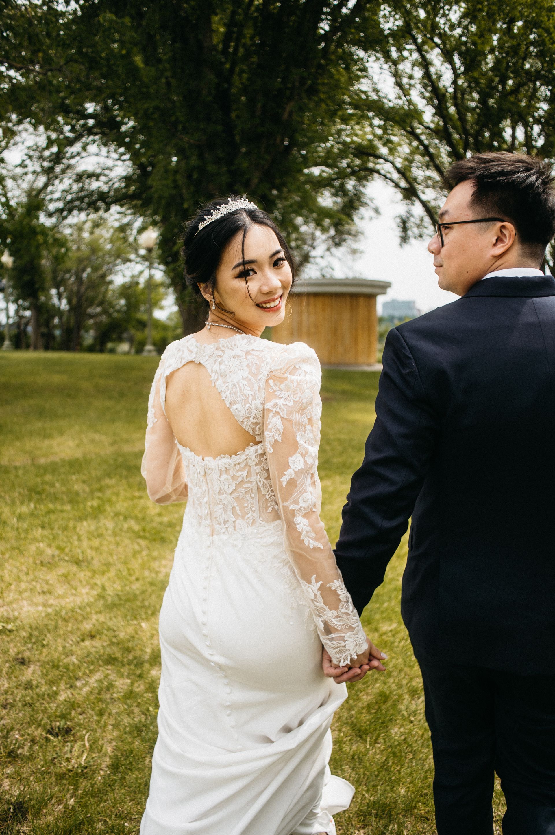 Bride in lace dress, looking back, holds hands with groom in navy suit, walking on grass.