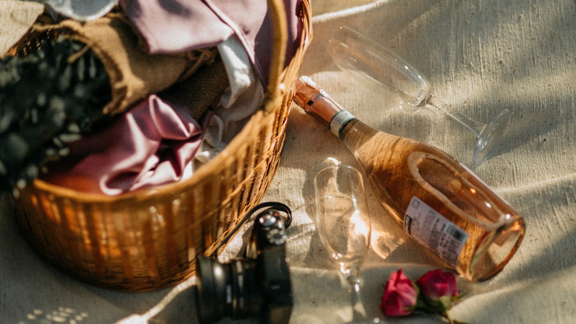 Picnic basket with pink wine bottle, champagne flutes, flowers, and cloth on a textured surface.