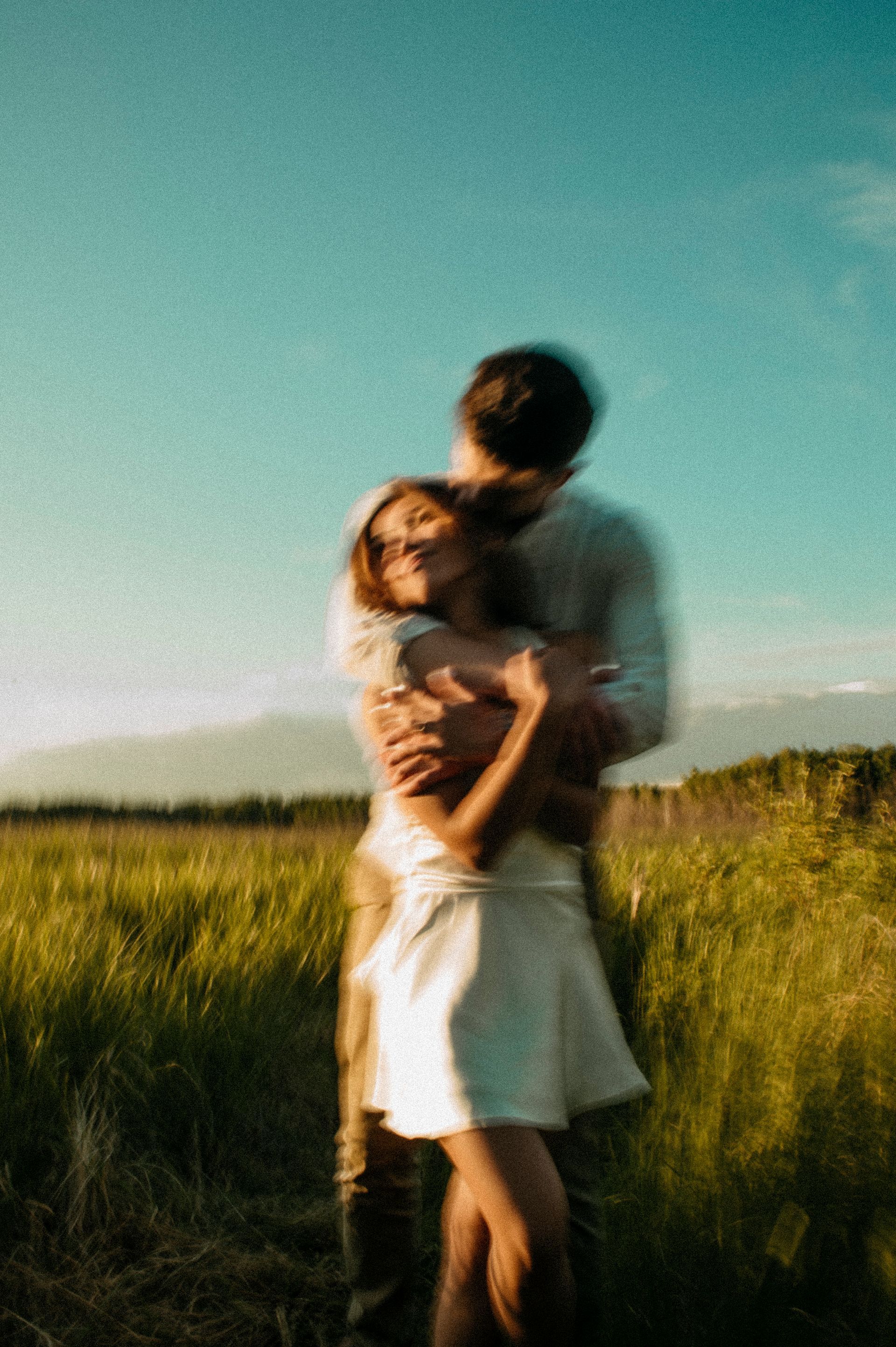 Couple hugging in a field of grass. She wears a white dress; he has his arms around her. Blue sky.