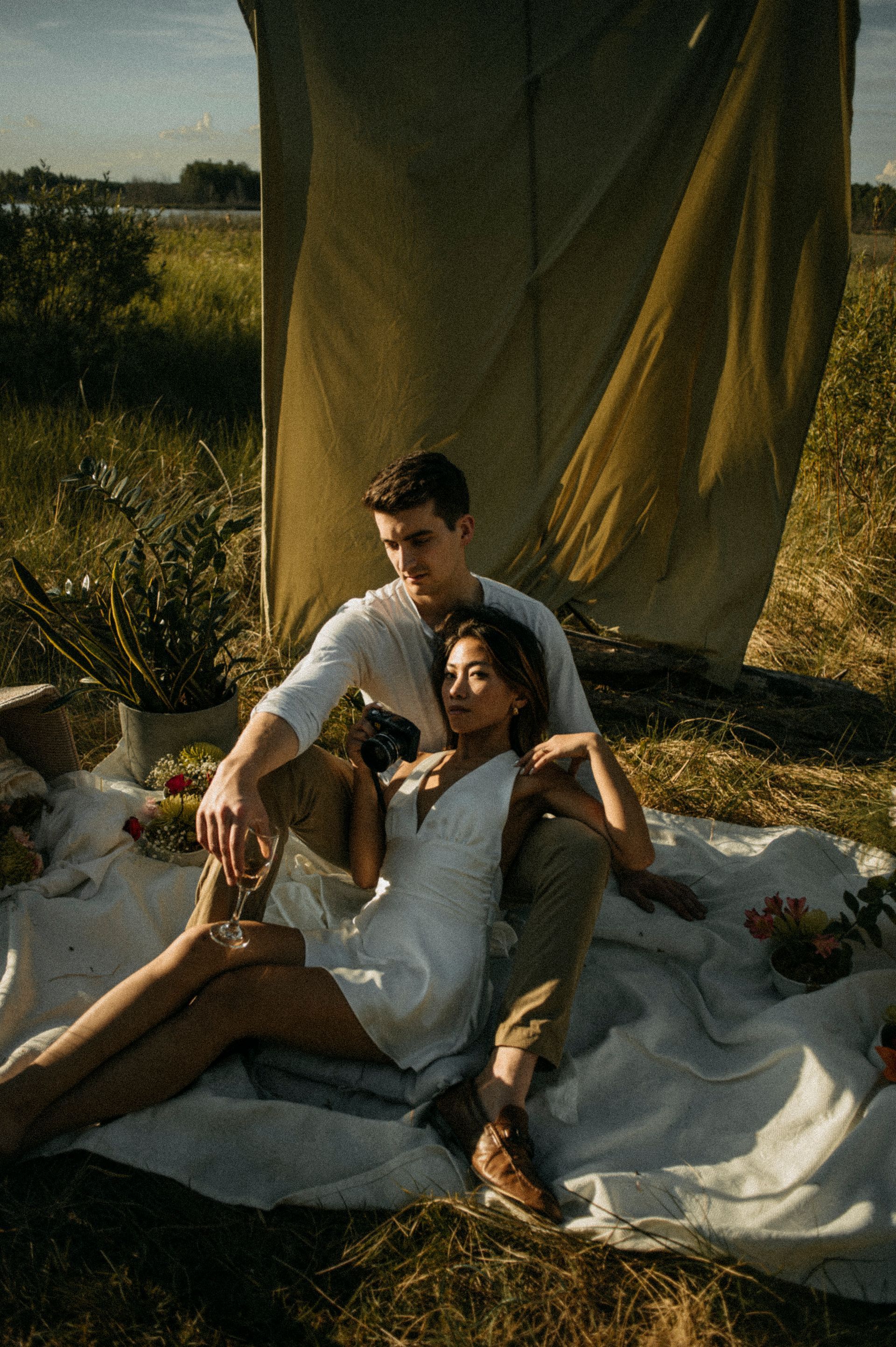 Couple sitting on a blanket outdoors. Woman in white dress, man in white shirt. Picnic setting.