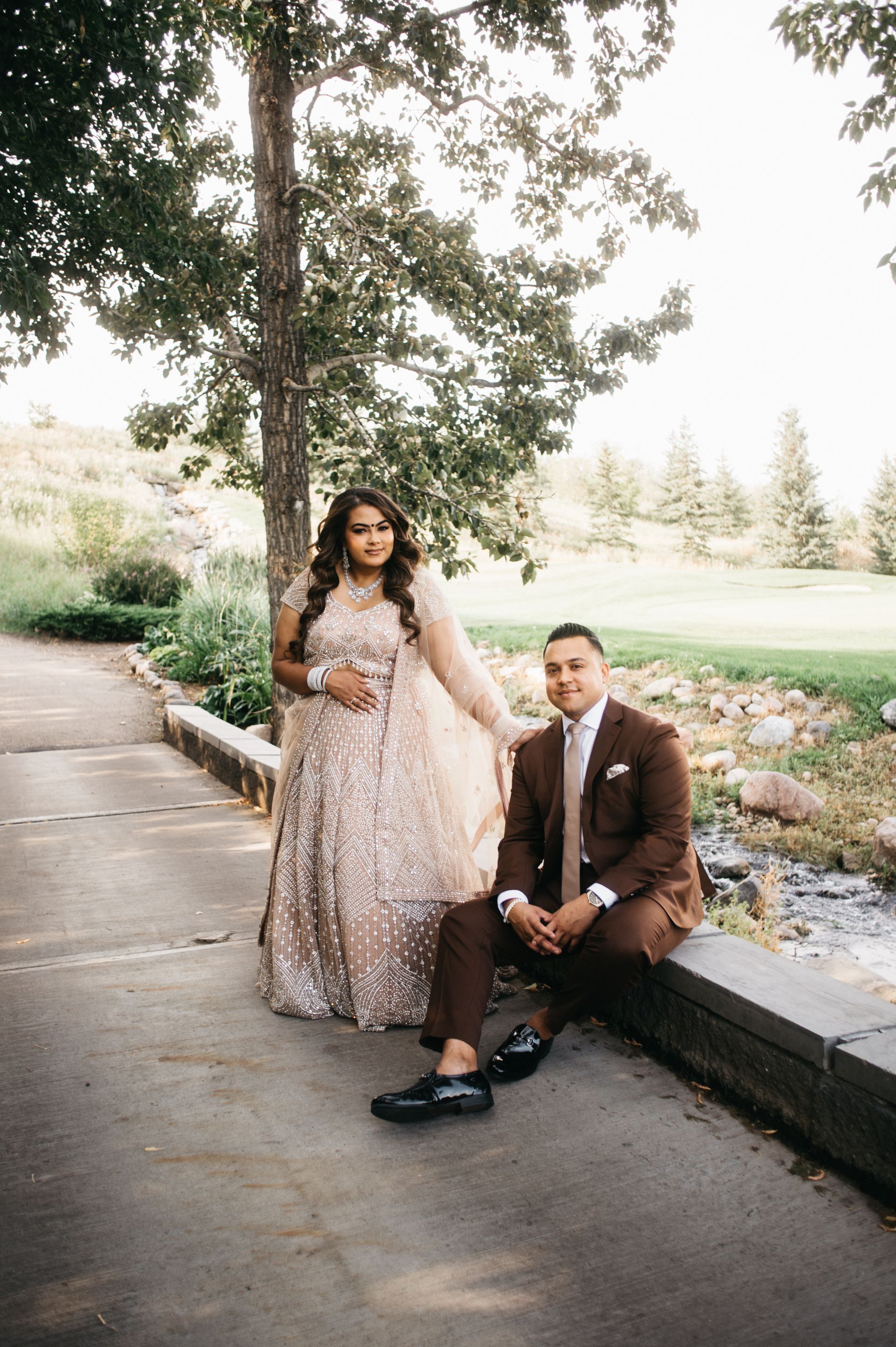 Couple in formal wear on a pathway. Man seated, looking at the camera. Woman stands, leaning toward man. Outdoors, near trees.