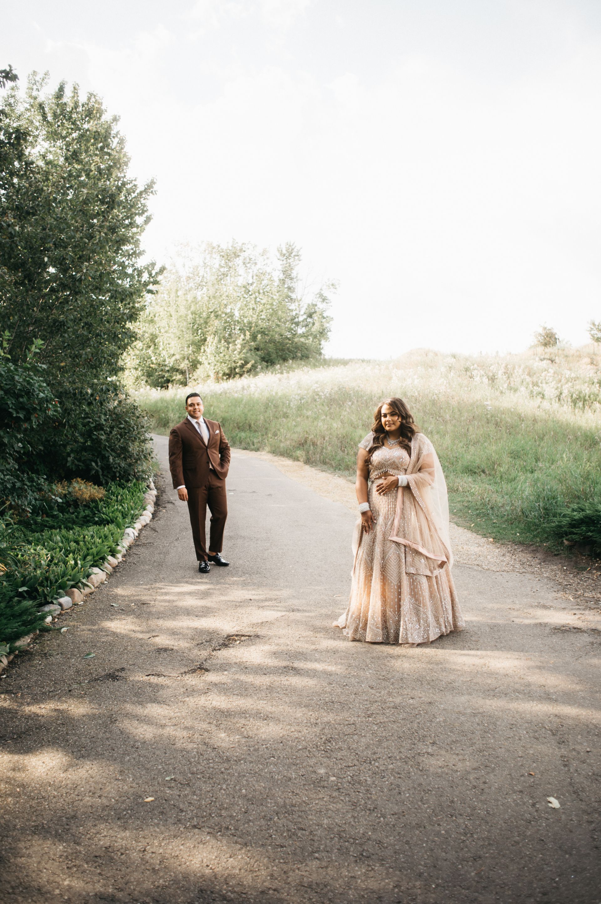 Couple walking on a paved path. Man in brown suit, woman in patterned gown; outdoor setting.