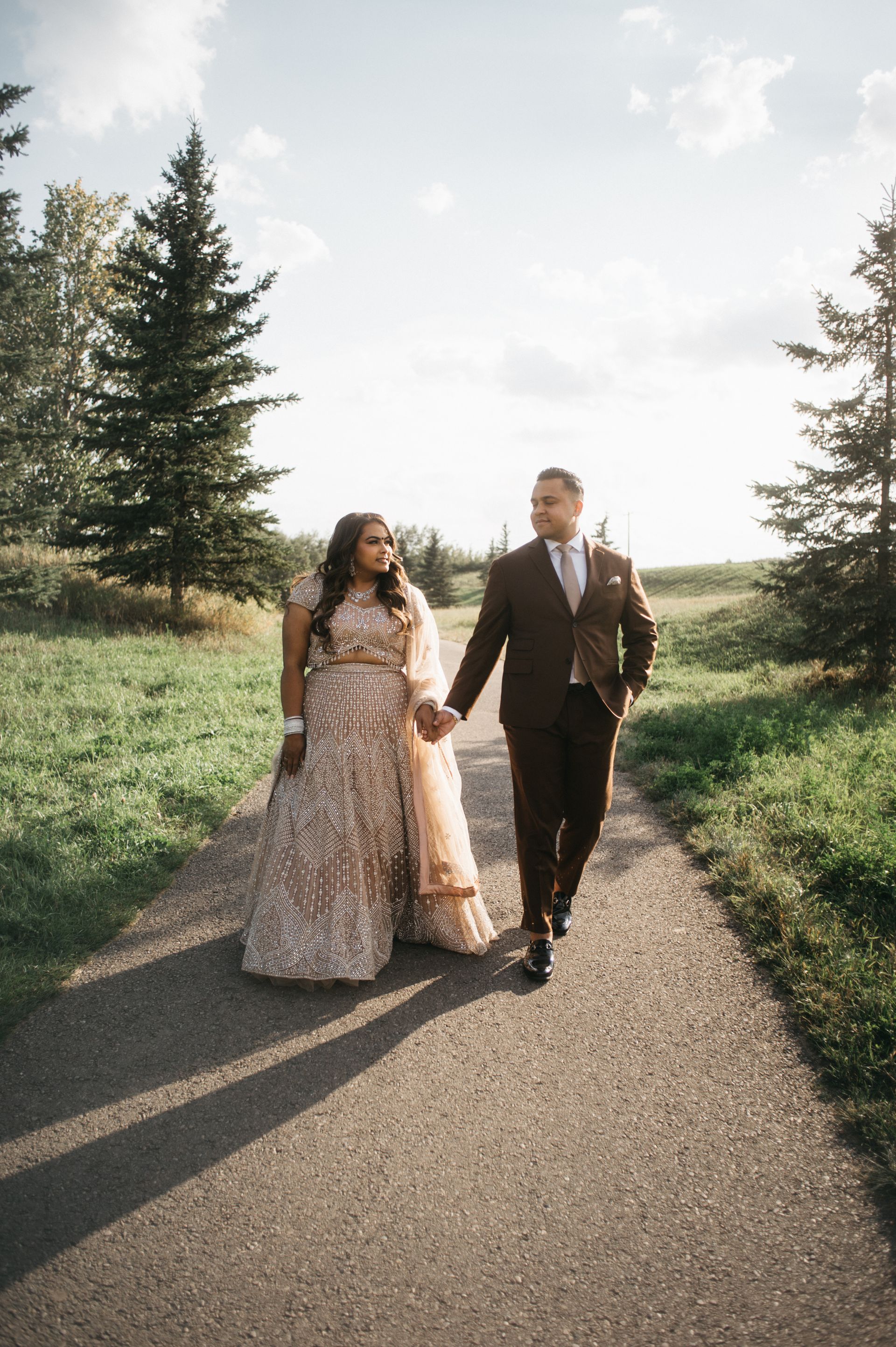 Couple in formal attire walk hand-in-hand down a path in a park, golden sunlight.