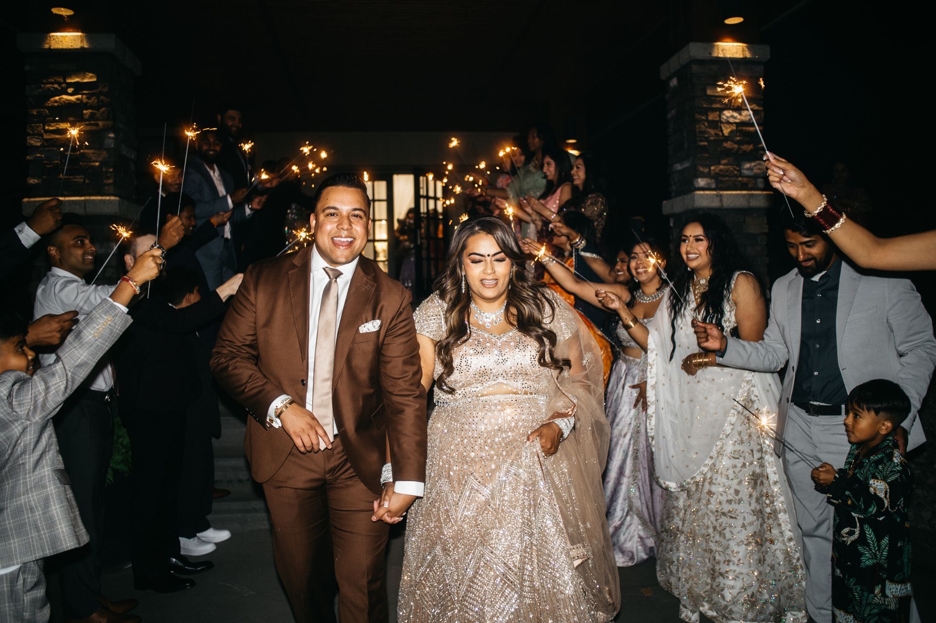 Couple holding hands, exiting a building at night, surrounded by sparklers held by guests.