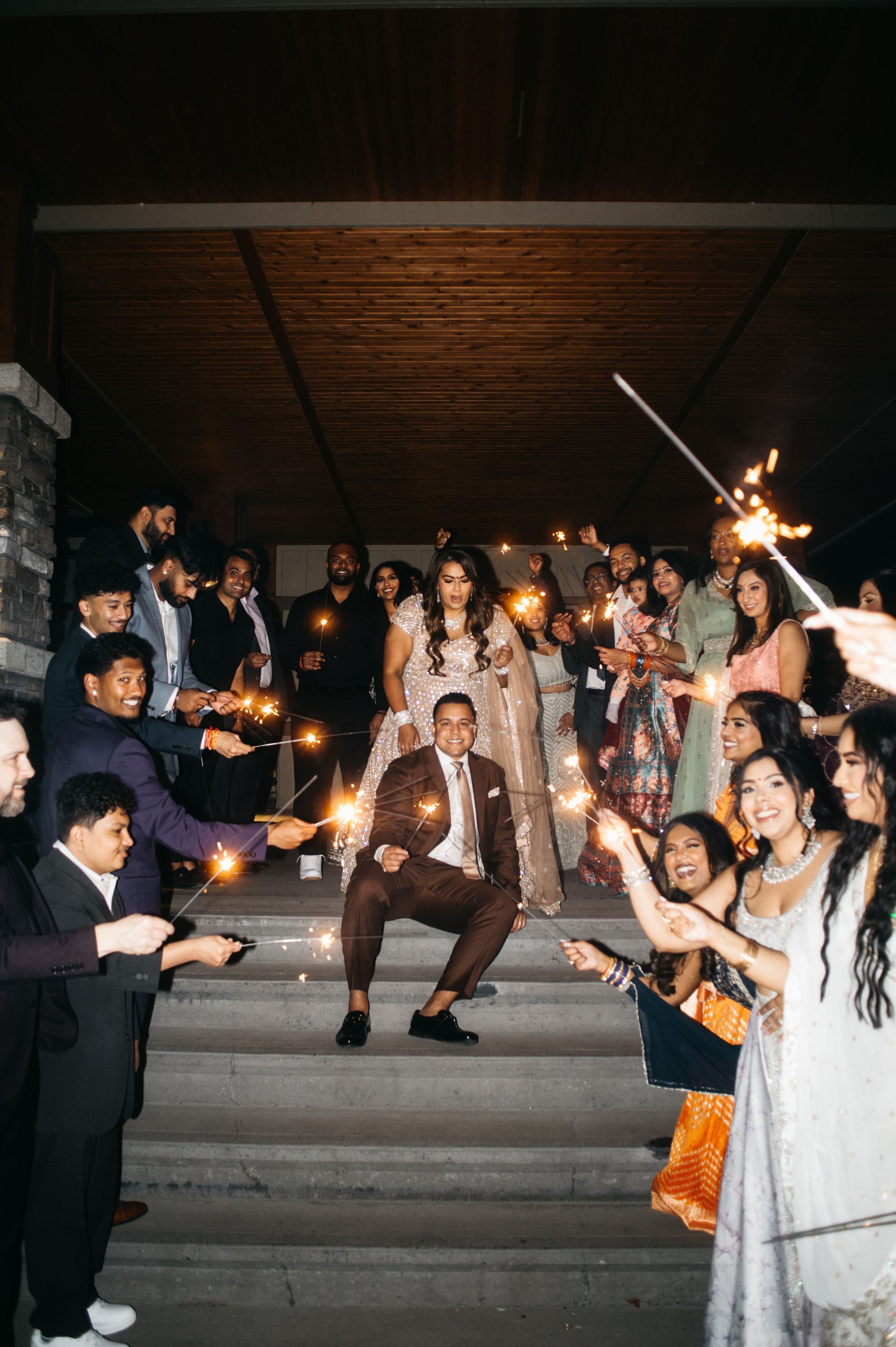 Couple crouches on steps, surrounded by guests holding sparklers, celebrating at night.