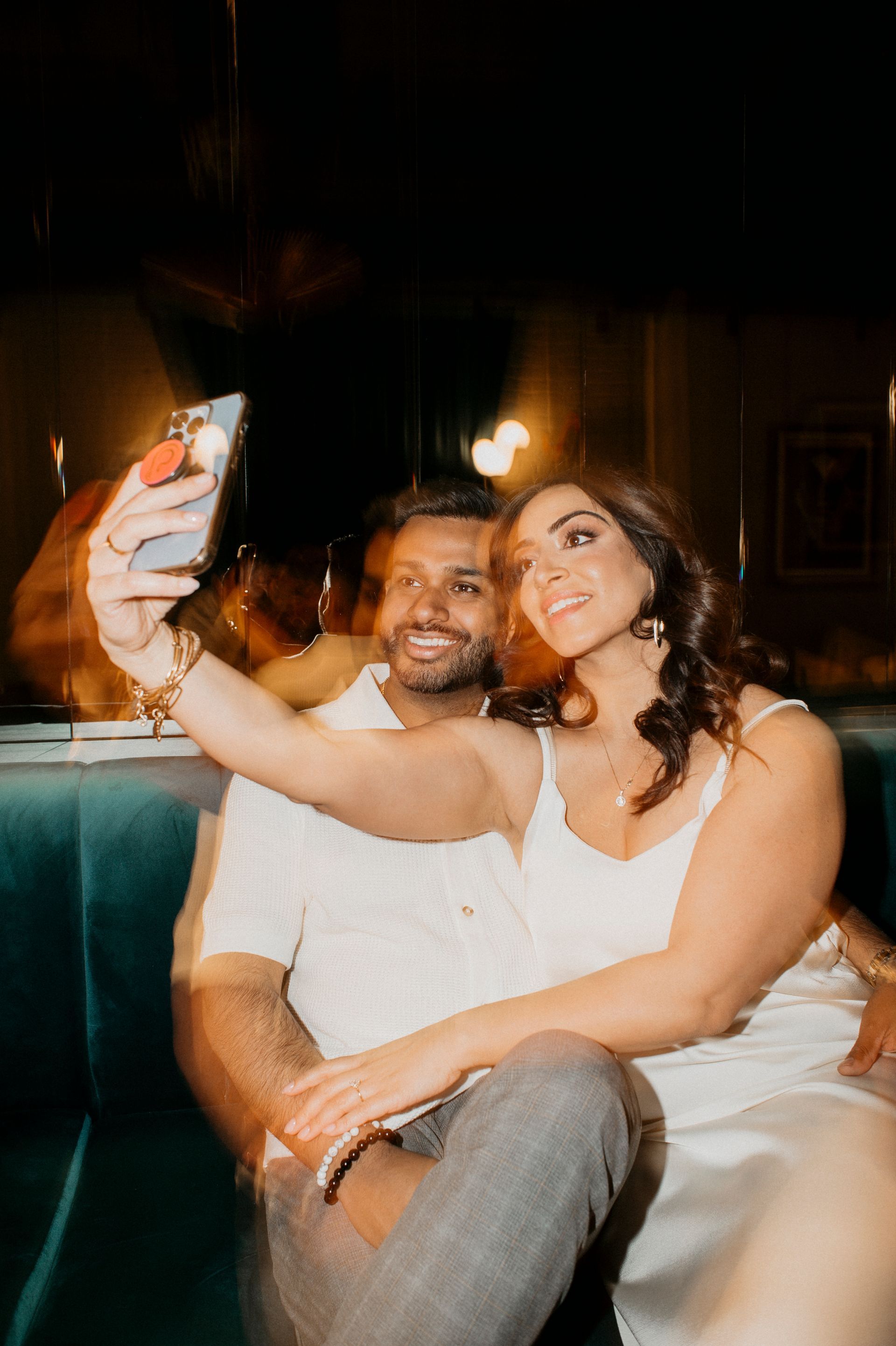 Couple on a teal couch, taking a selfie. Man smiling with arm around woman, who is smiling, holding phone. Dim, elegant setting.