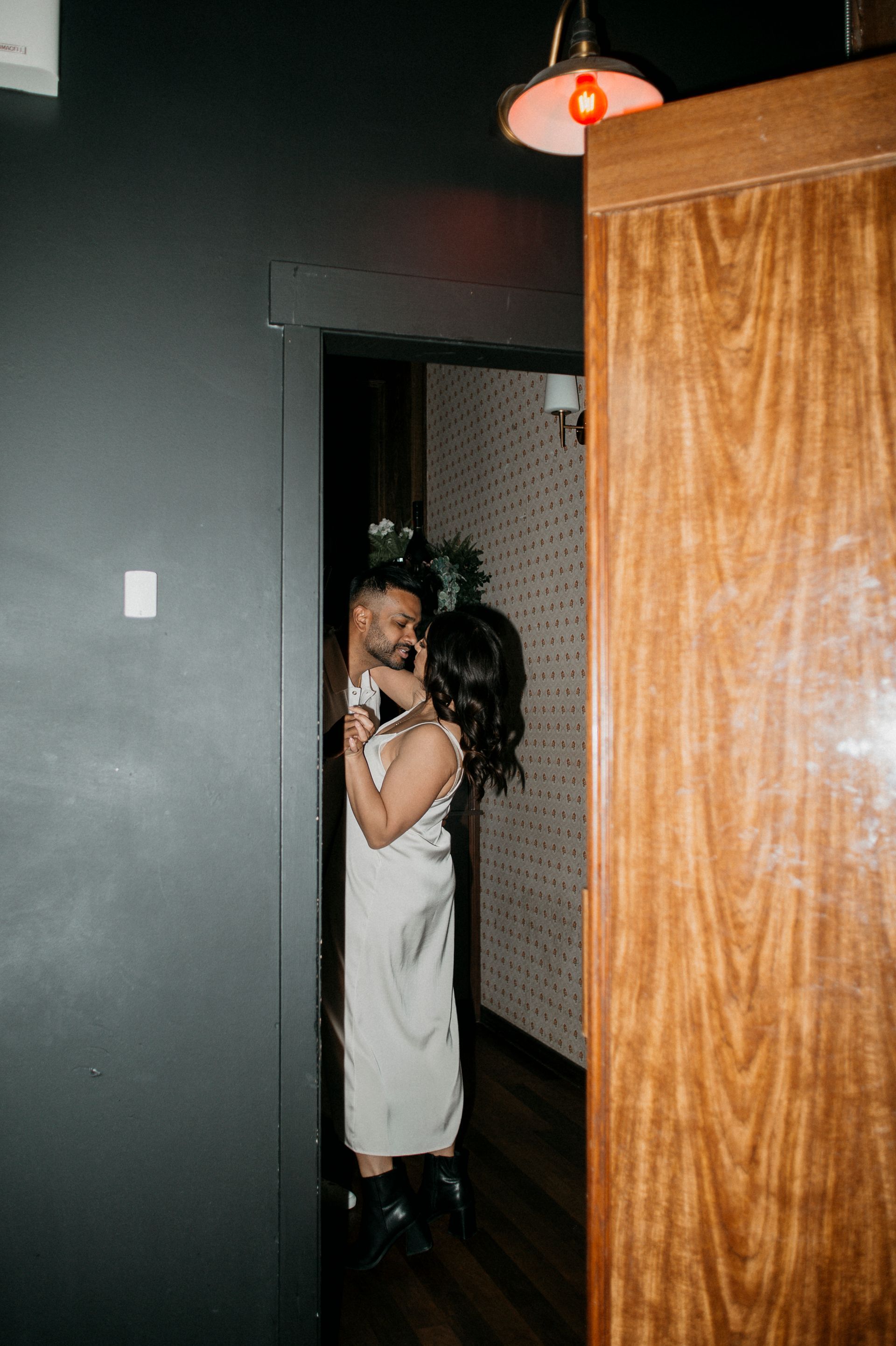 Couple embraces in a doorway, dimly lit. One wears a satin dress, other in dark clothing. Wooden door frame.
