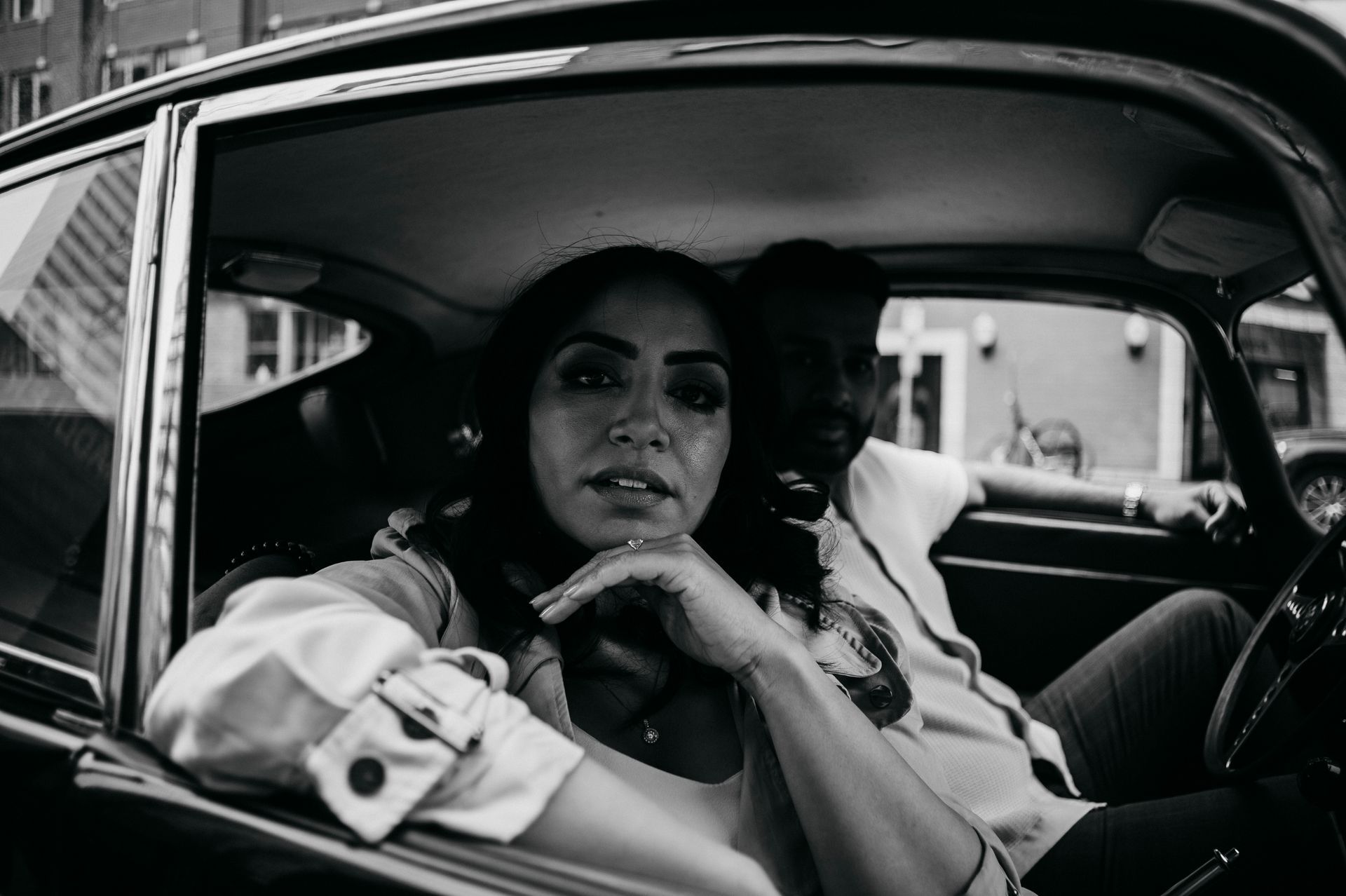 Woman in classic car, looking towards camera, arm raised. Another person partially visible in the back seat. Black and white.