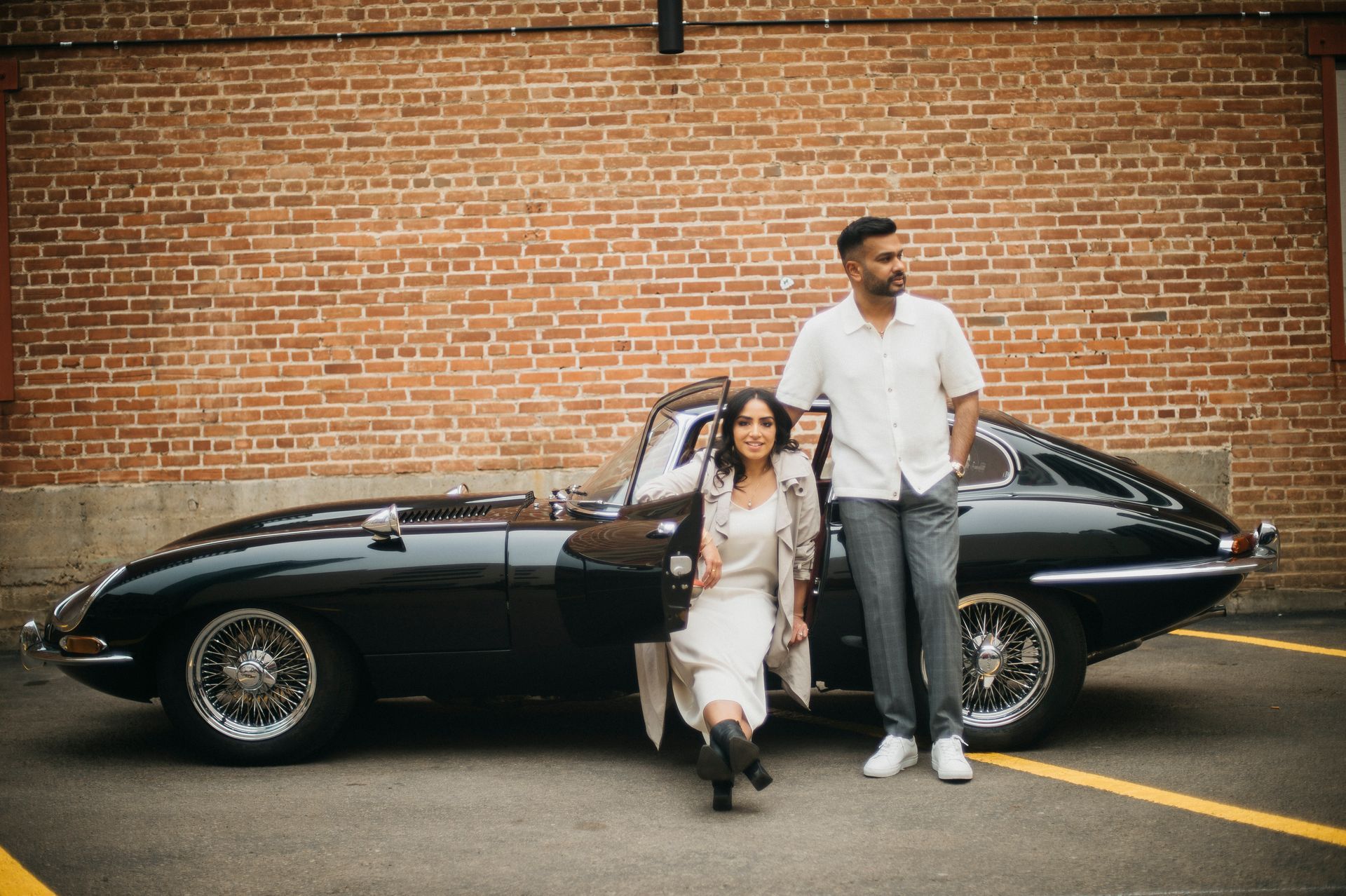Couple posing with a vintage black car against a brick wall.