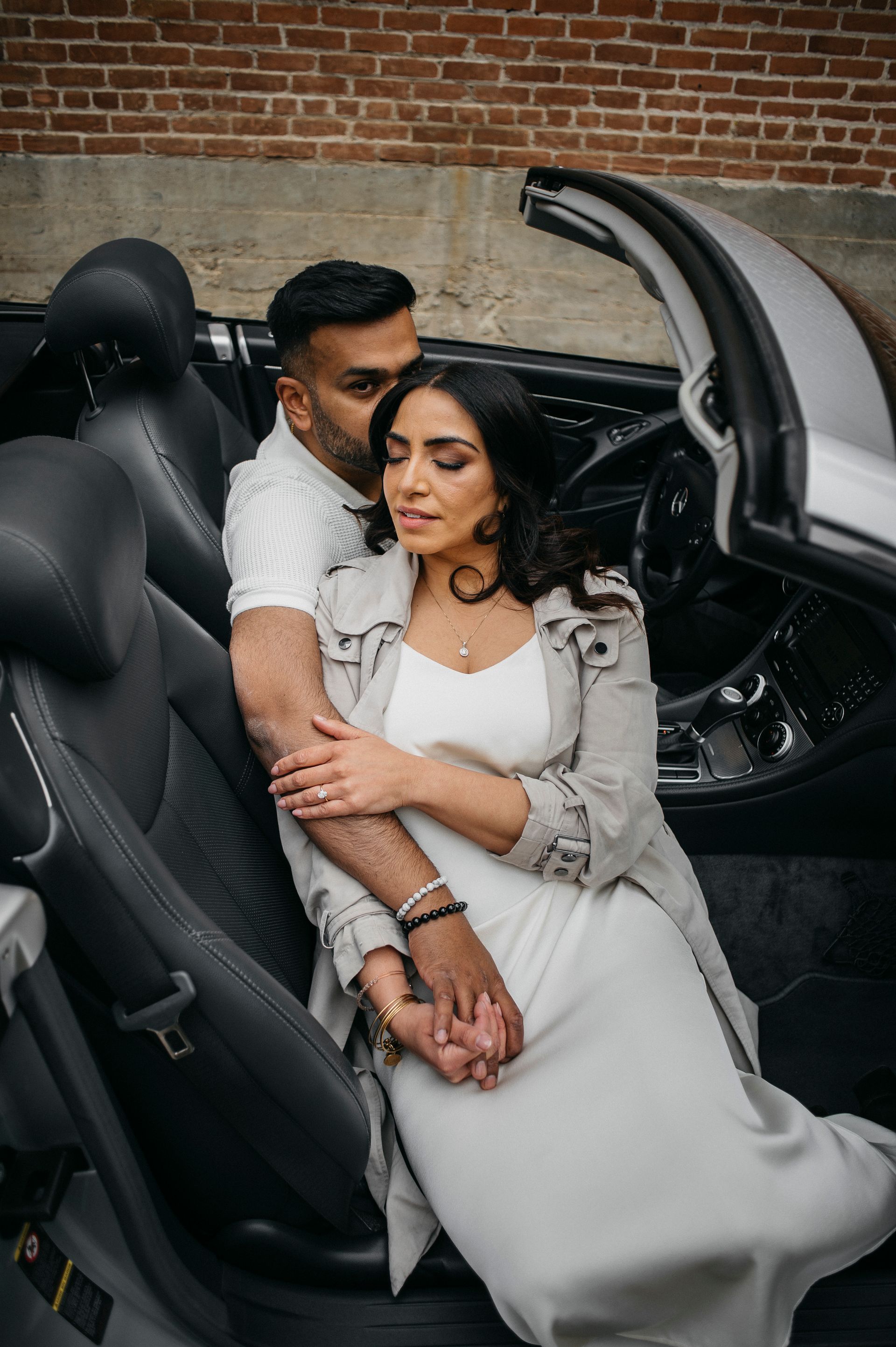 Couple in convertible, man embracing woman. Car interior is black, they are by brick wall.