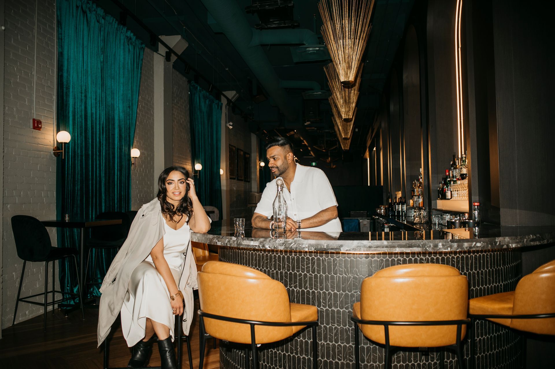 Woman and man at a bar, woman seated, man behind the bar. Teal curtains, gold fixtures, and yellow chairs.