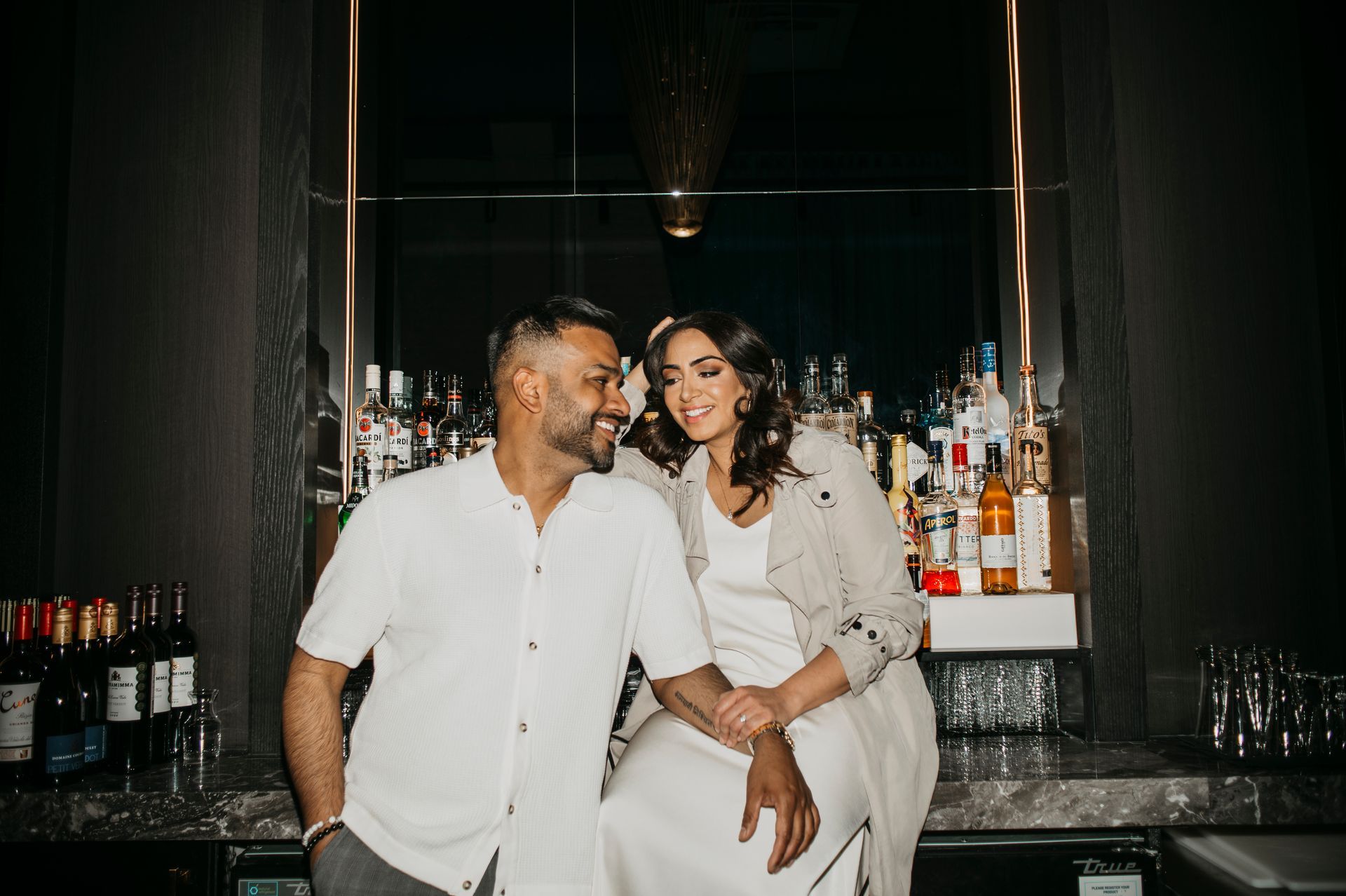 Couple smiling at each other, sitting on a bar, surrounded by bottles.