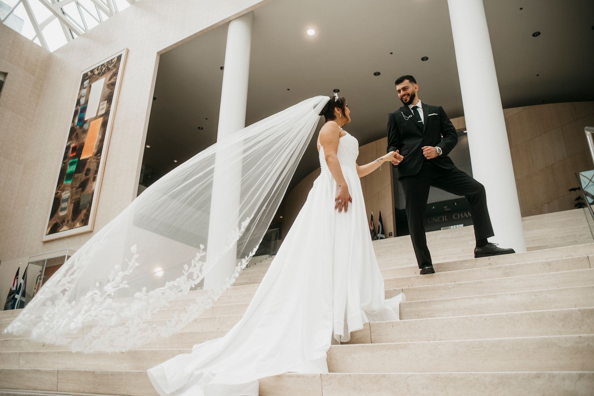 Bride and groom on stairs, bride in a white dress with veil, groom in black suit, holding hands, modern building interior.
