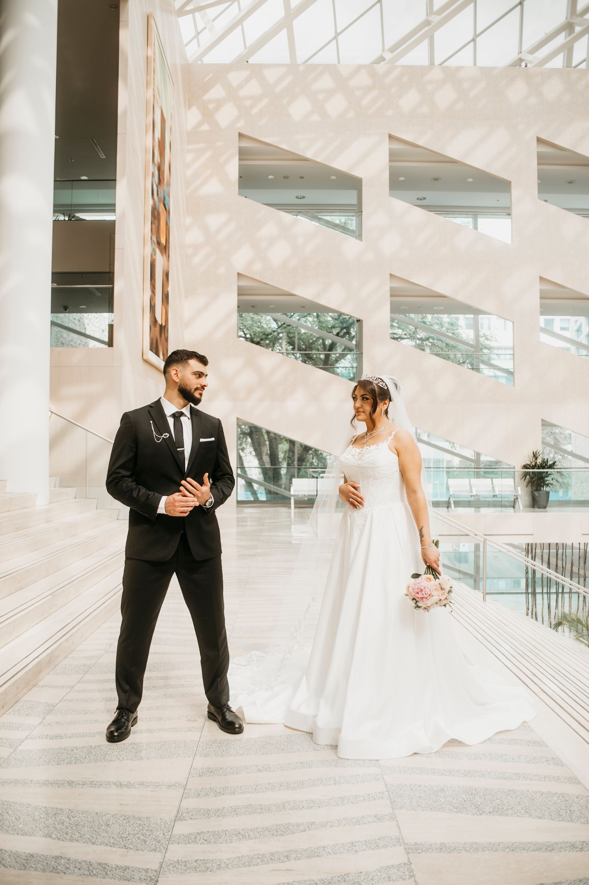 Bride and groom in wedding attire pose indoors. The bride holds a bouquet, the groom looks at her. Modern building backdrop.