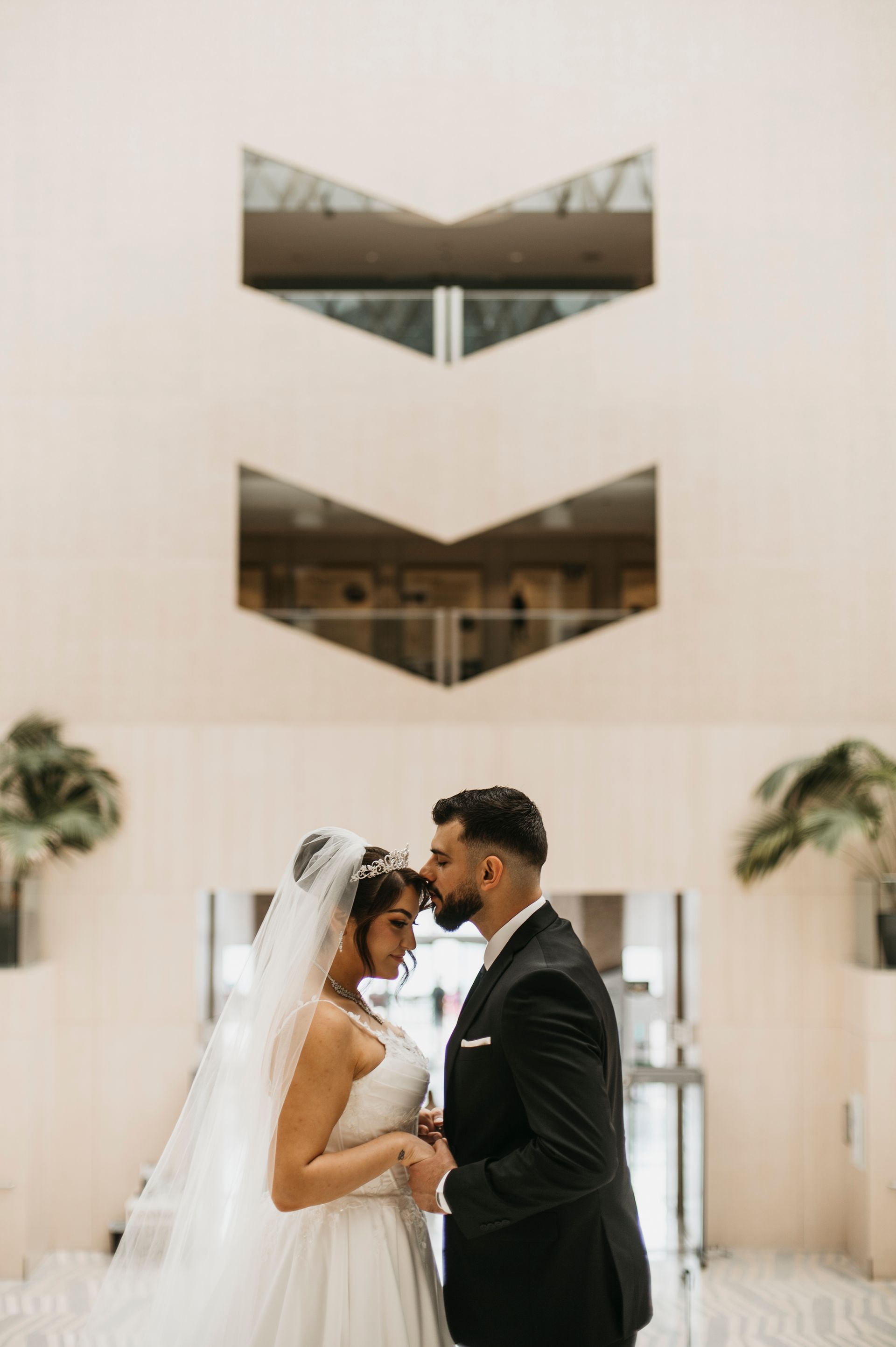 Bride and groom in wedding attire touch foreheads in front of a beige building with geometric windows.