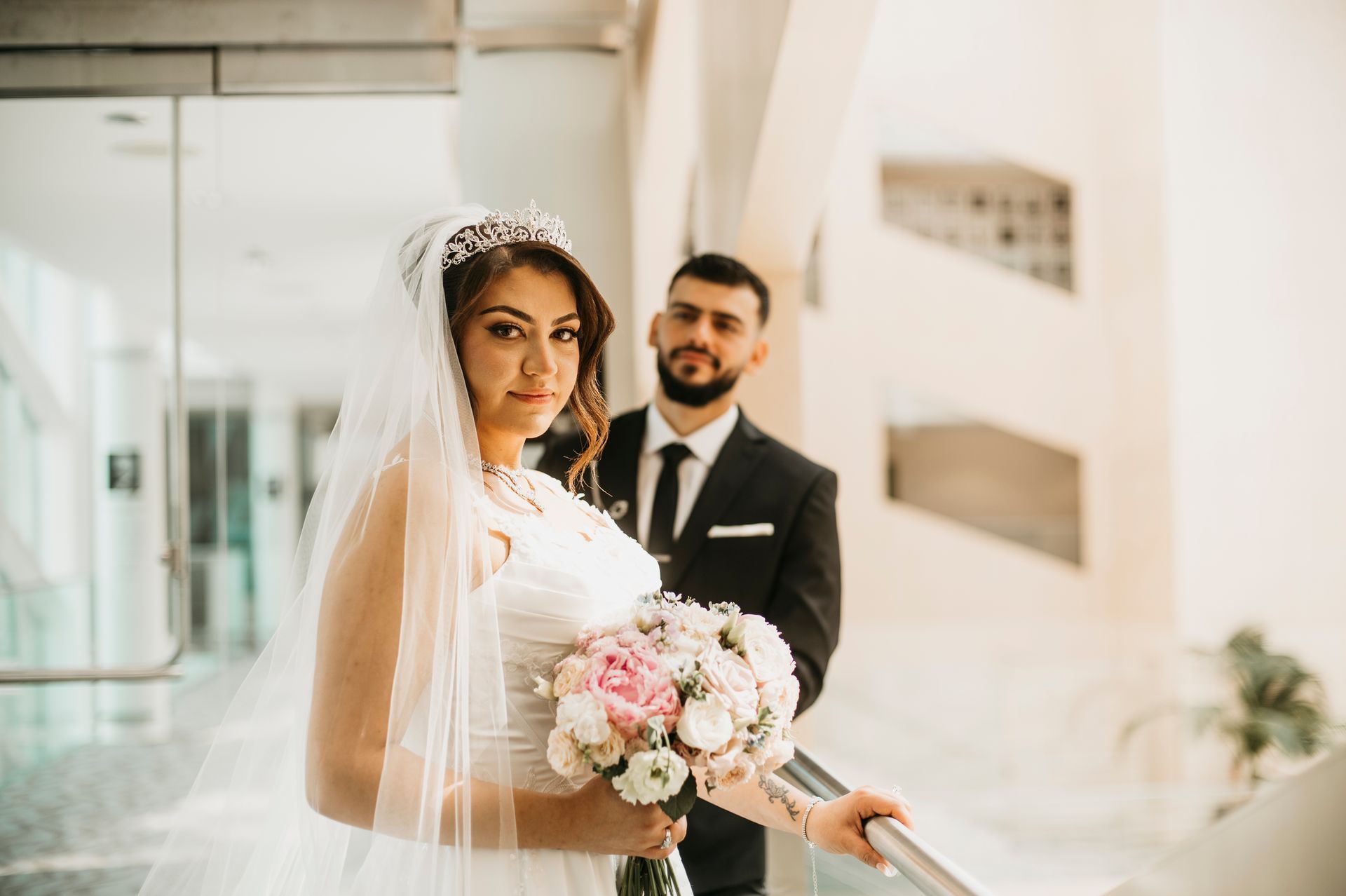 Bride in white gown, veil, and tiara, holding flowers. Groom in suit stands behind her in a building.