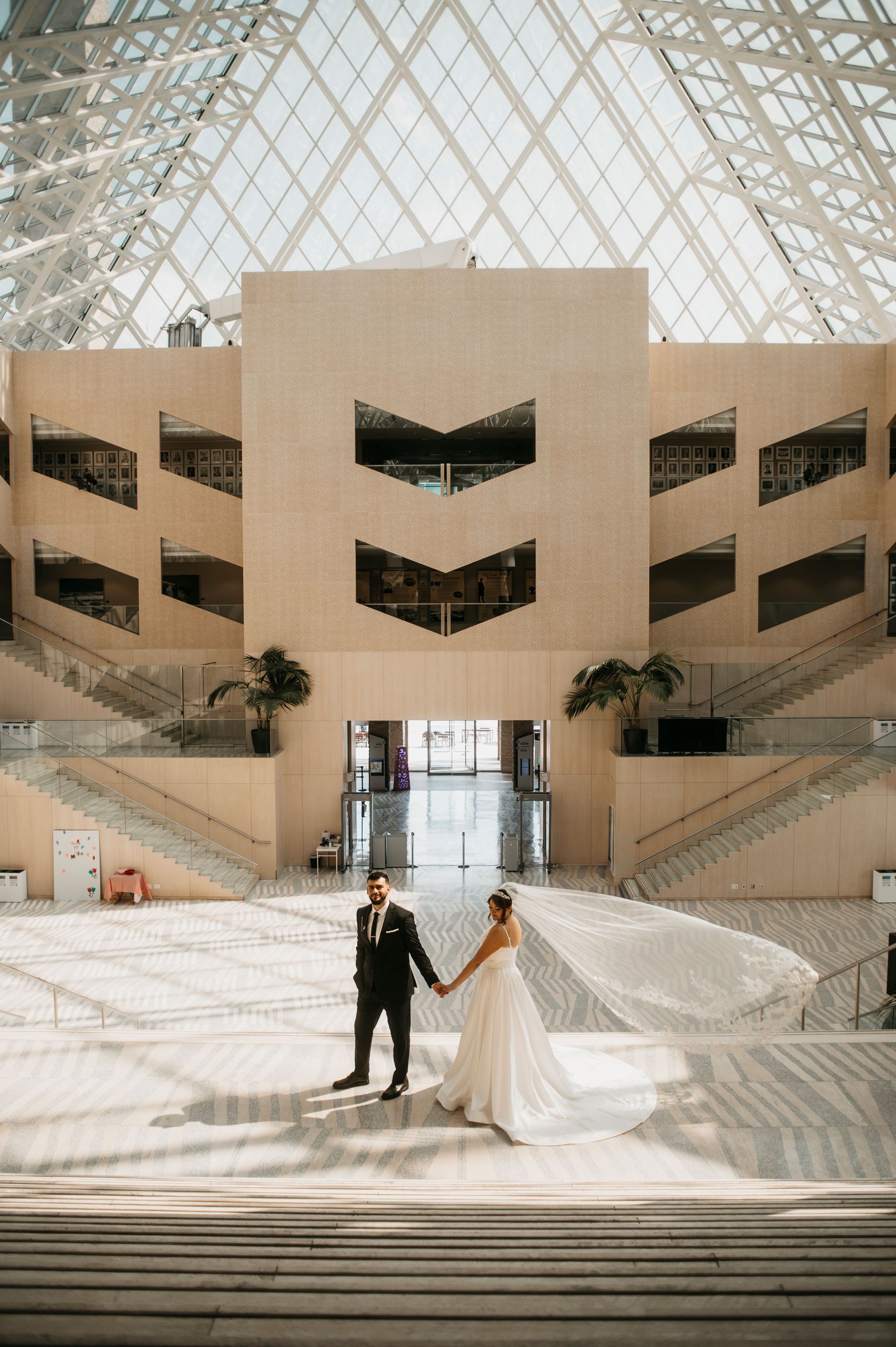 Newlyweds holding hands pose in a modern building with a glass ceiling. The bride's veil flows.