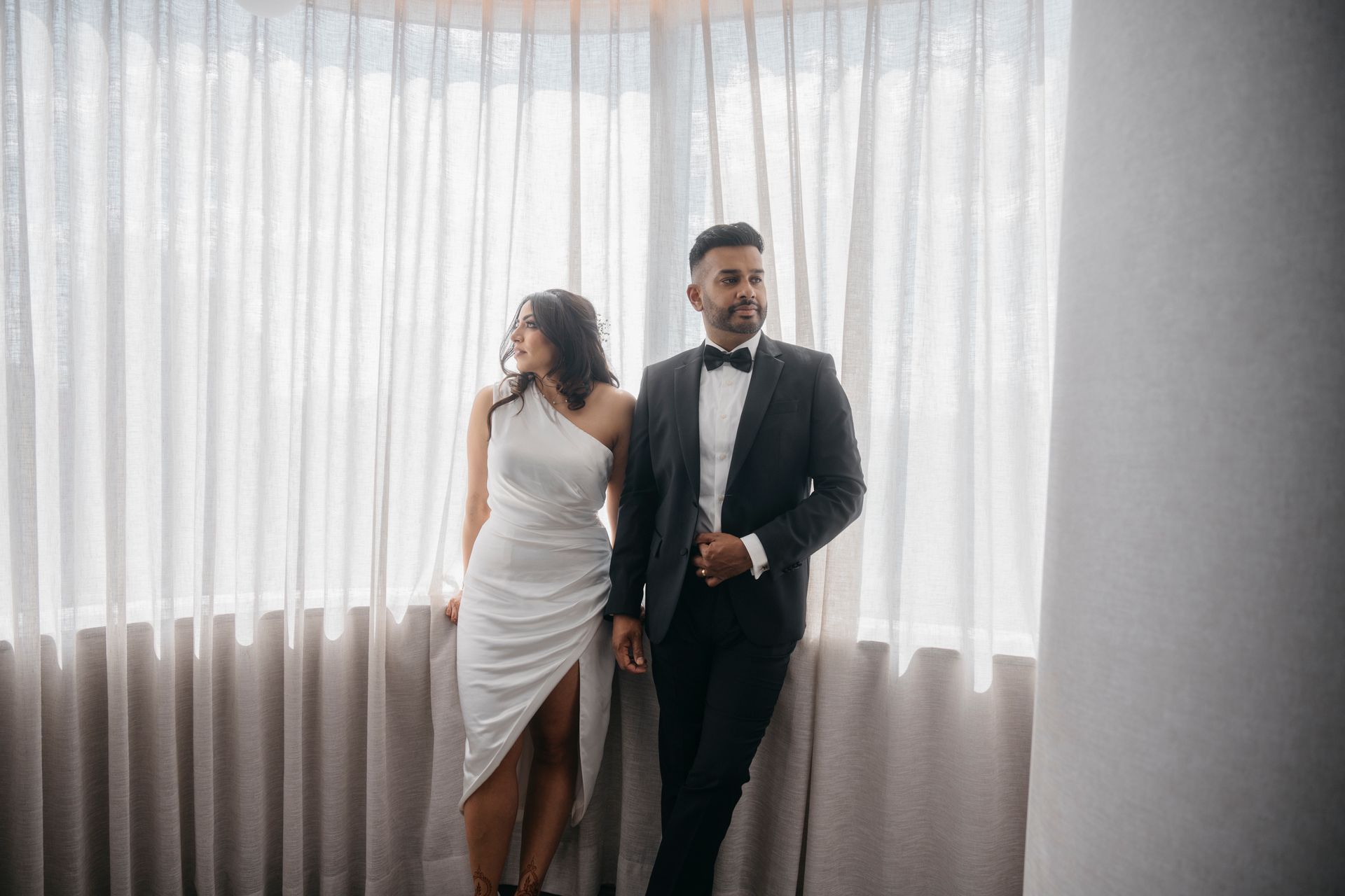 Couple in formal attire by a window; woman in white dress, man in black suit, looking away.