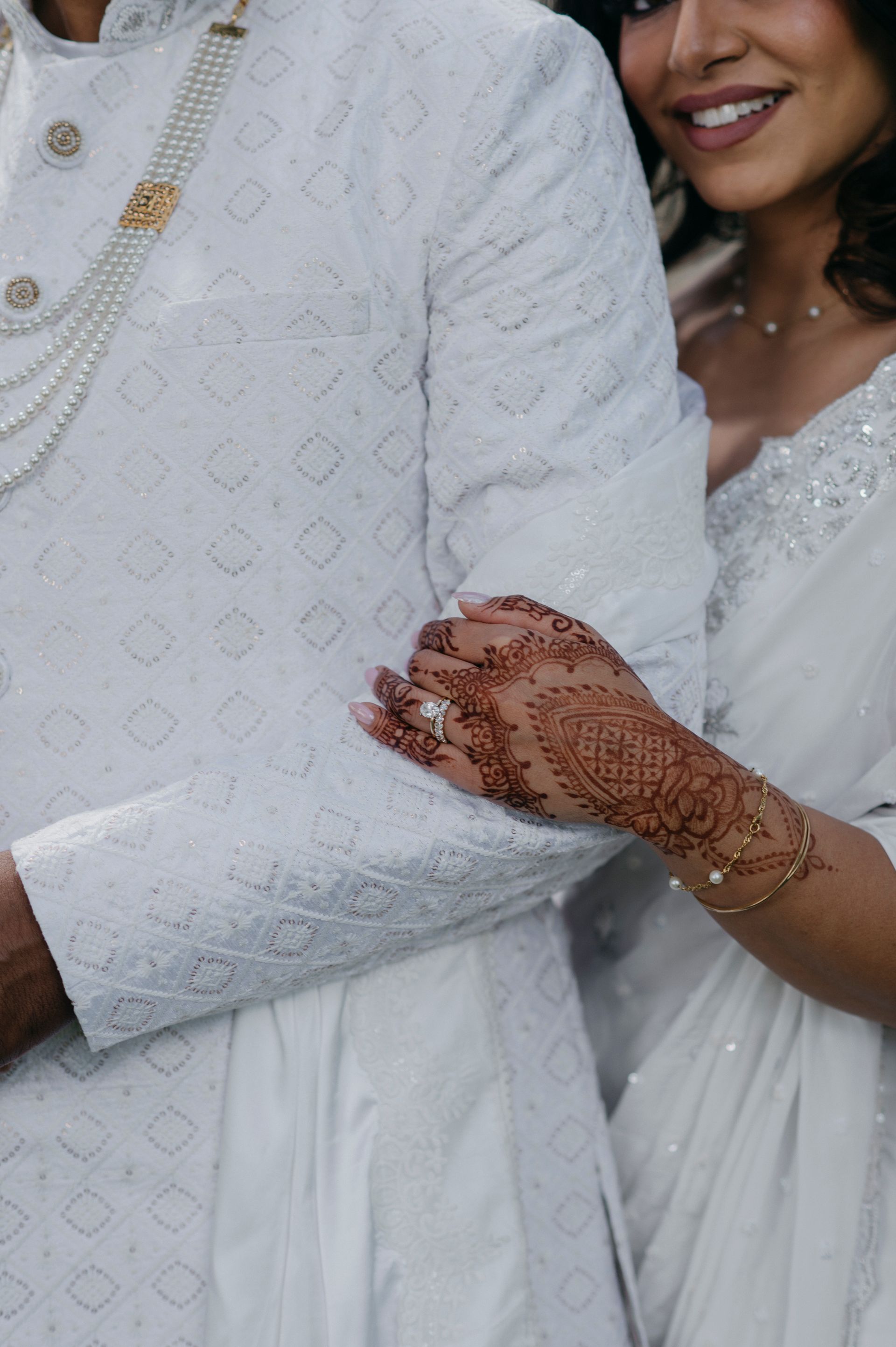 Couple in white outfits embrace; woman's hand with henna on man's arm.