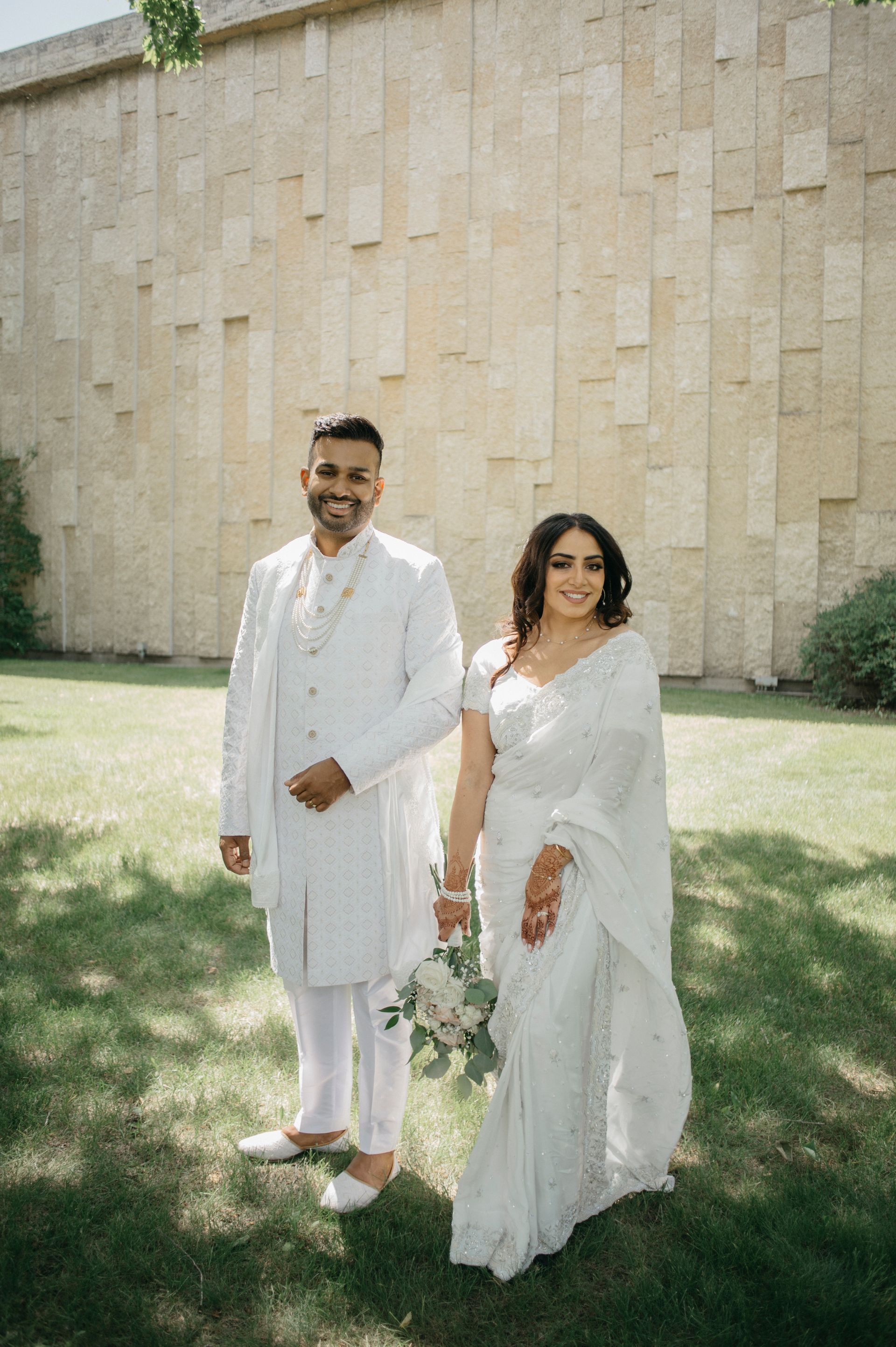 Couple in white wedding attire standing on grass, smiling. Building in the background.