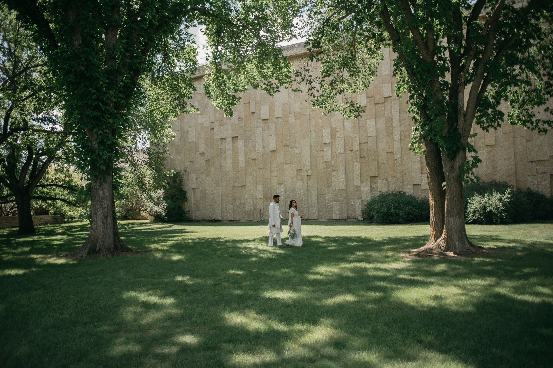 Couple standing in front of a textured wall, beneath the shade of trees on a grassy lawn.