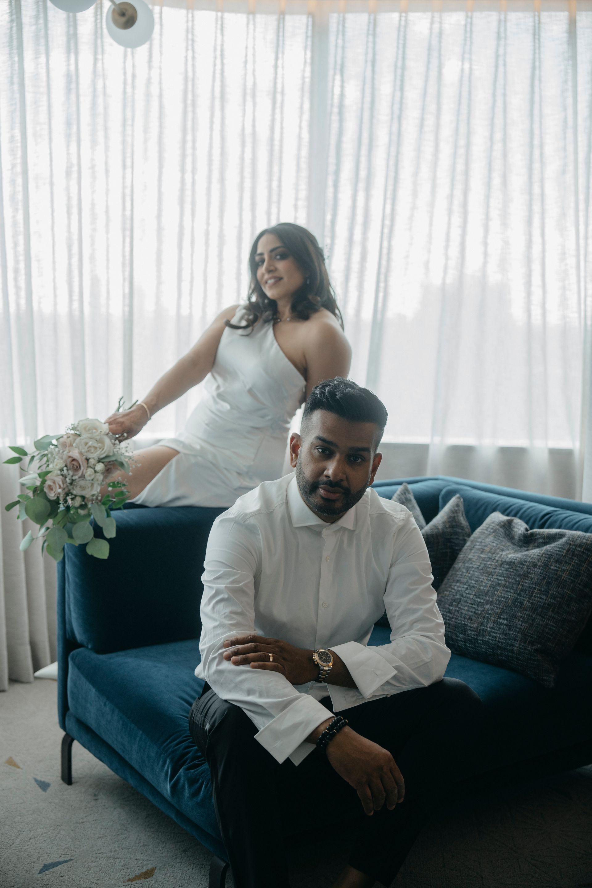 Couple posing; woman in white dress on blue couch, man in white shirt, neutral background.