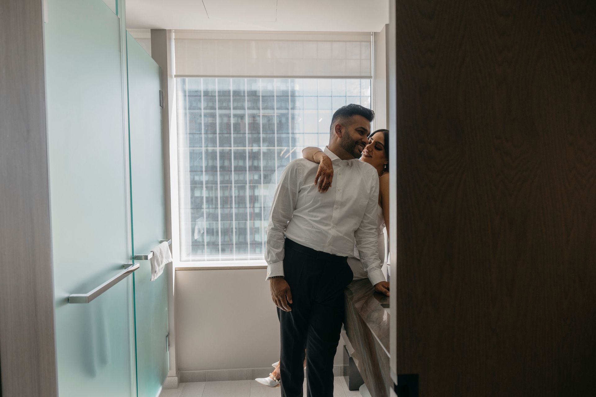 Couple embraces near a window. Man in white shirt, black pants; woman in light dress. Indoor setting.
