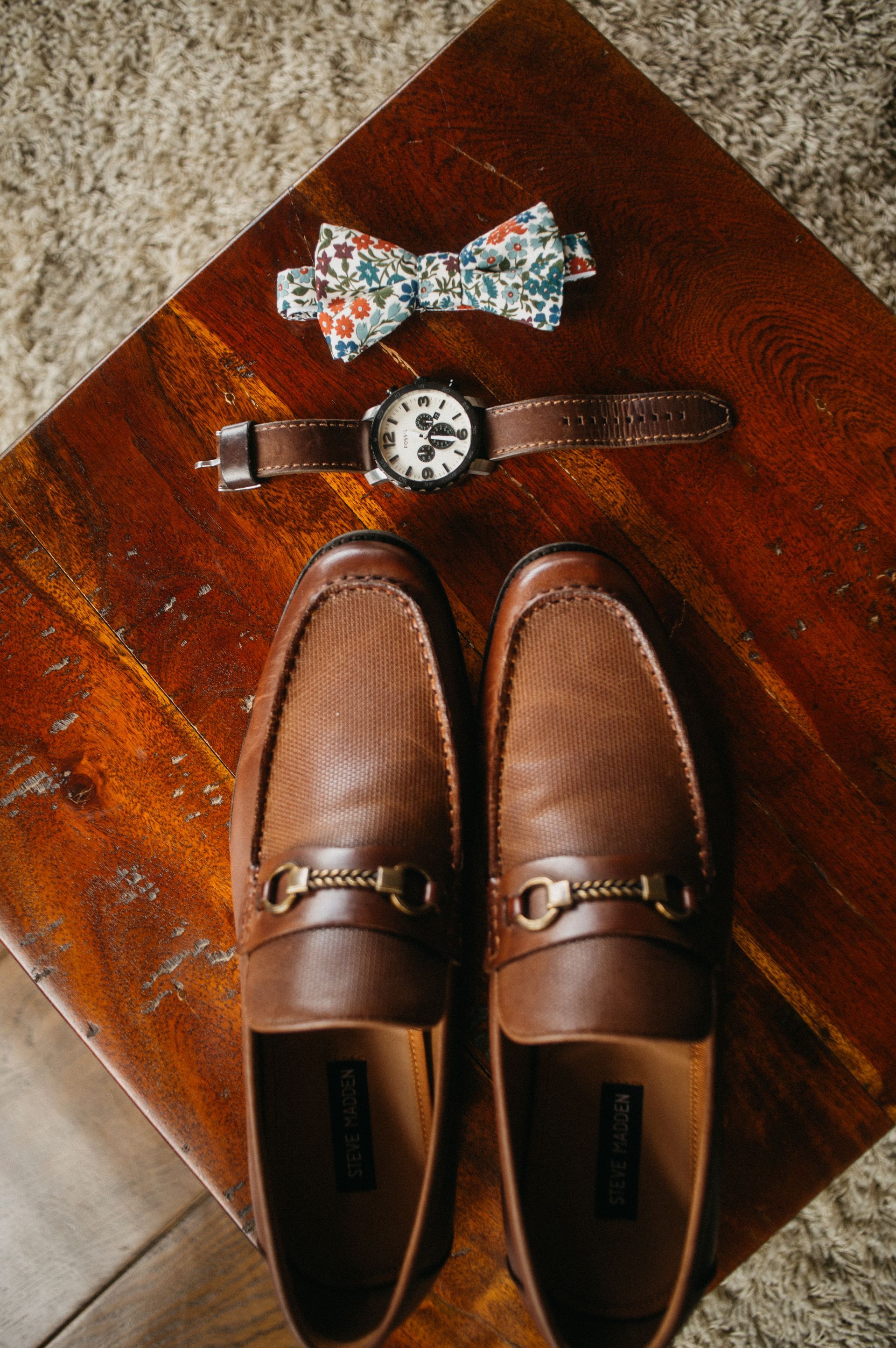 Brown loafers, watch, and patterned bow tie on a wooden surface.