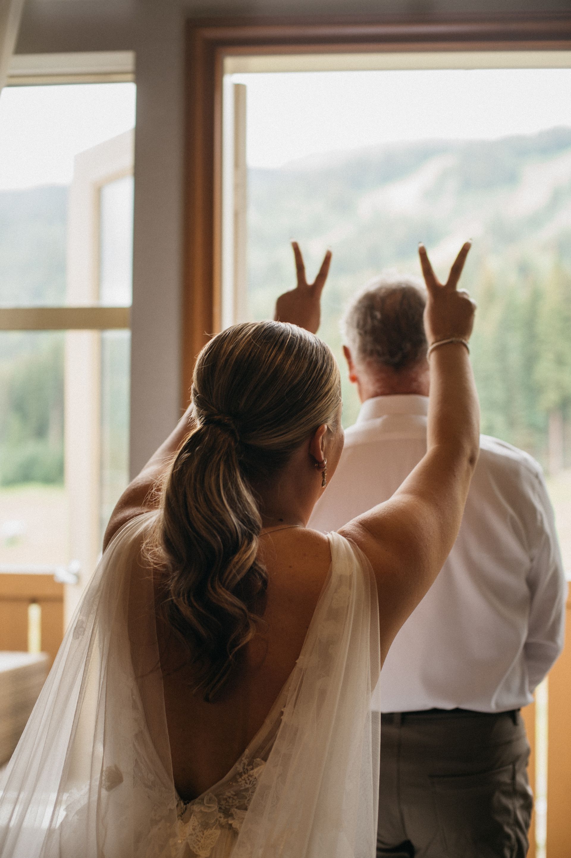 Bride with upraised arms making peace signs, standing near a window. Man in background.