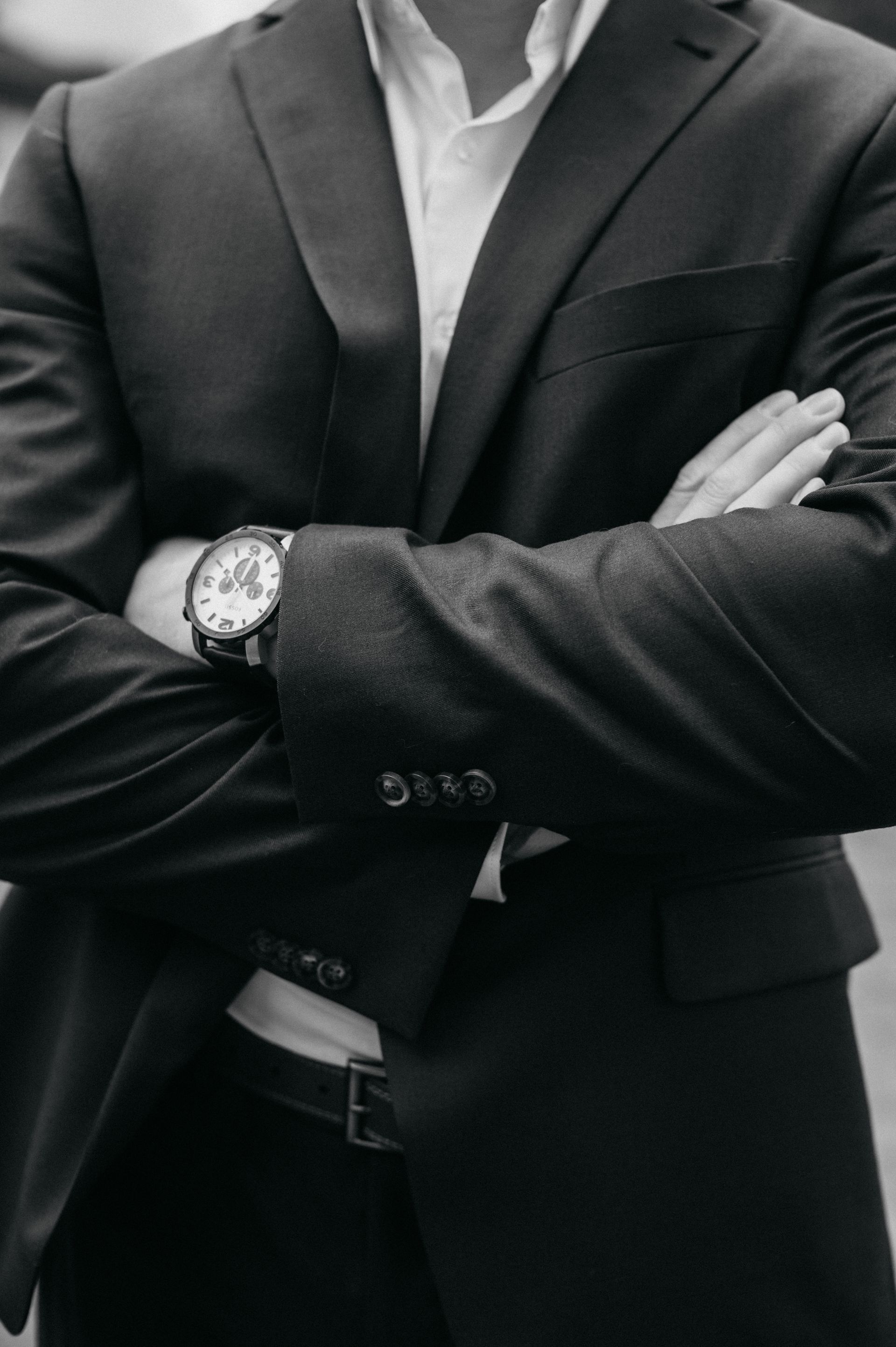Man in dark suit, arms crossed, wearing a wristwatch.