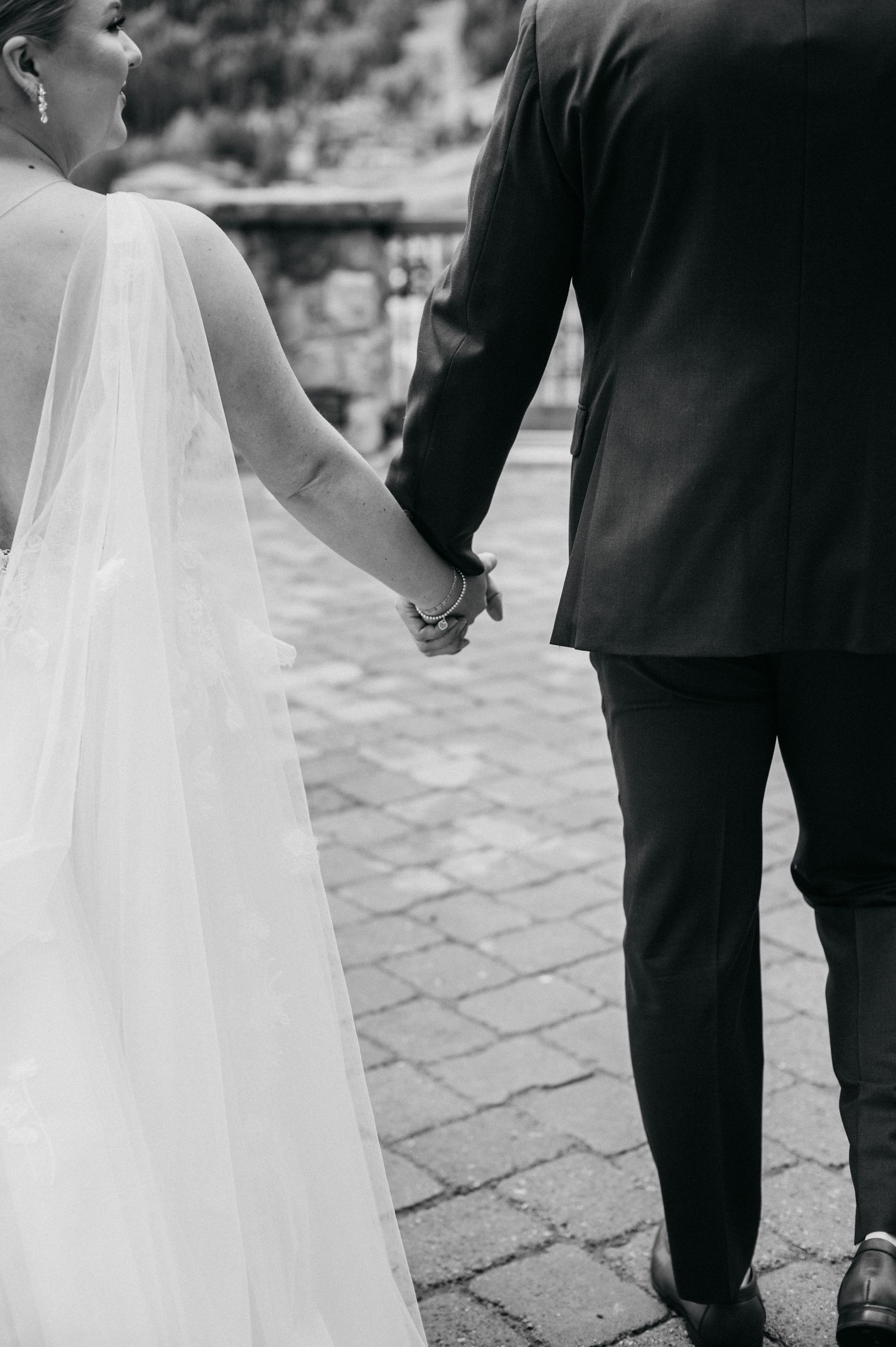Bride and groom holding hands, walking on a brick pathway, back view, black and white.