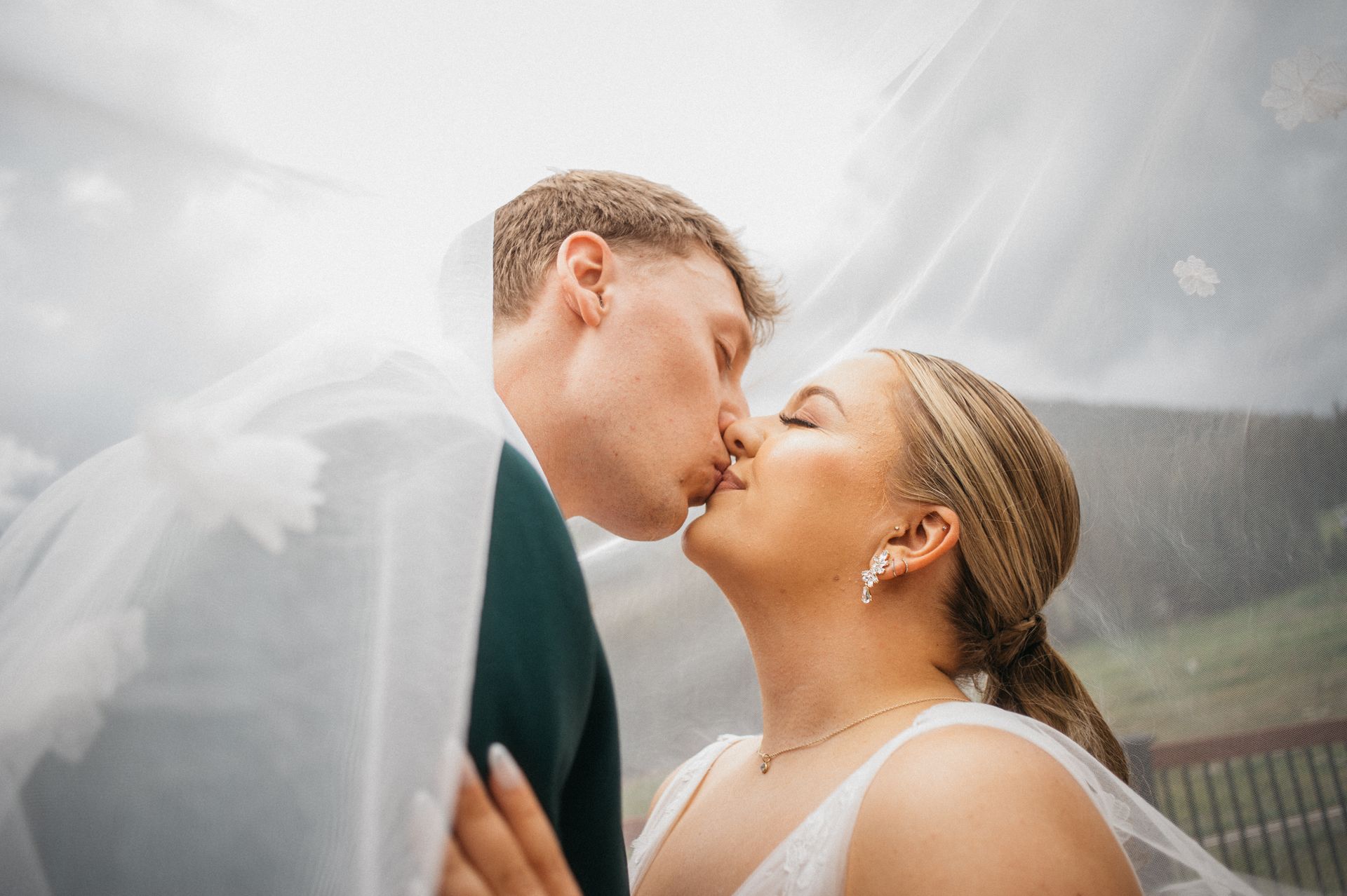 Couple kissing under a wedding veil; cloudy sky in the background.