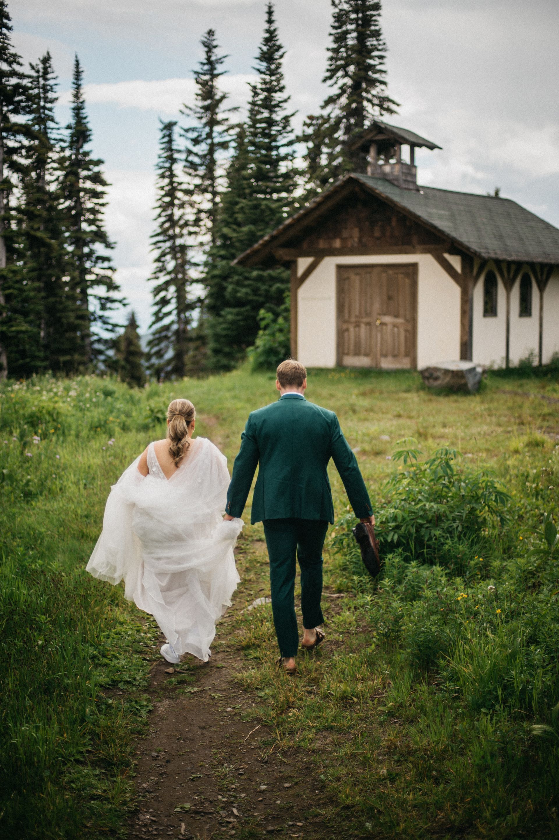Bride and groom walk hand-in-hand on a path towards a small chapel, mountains in background.