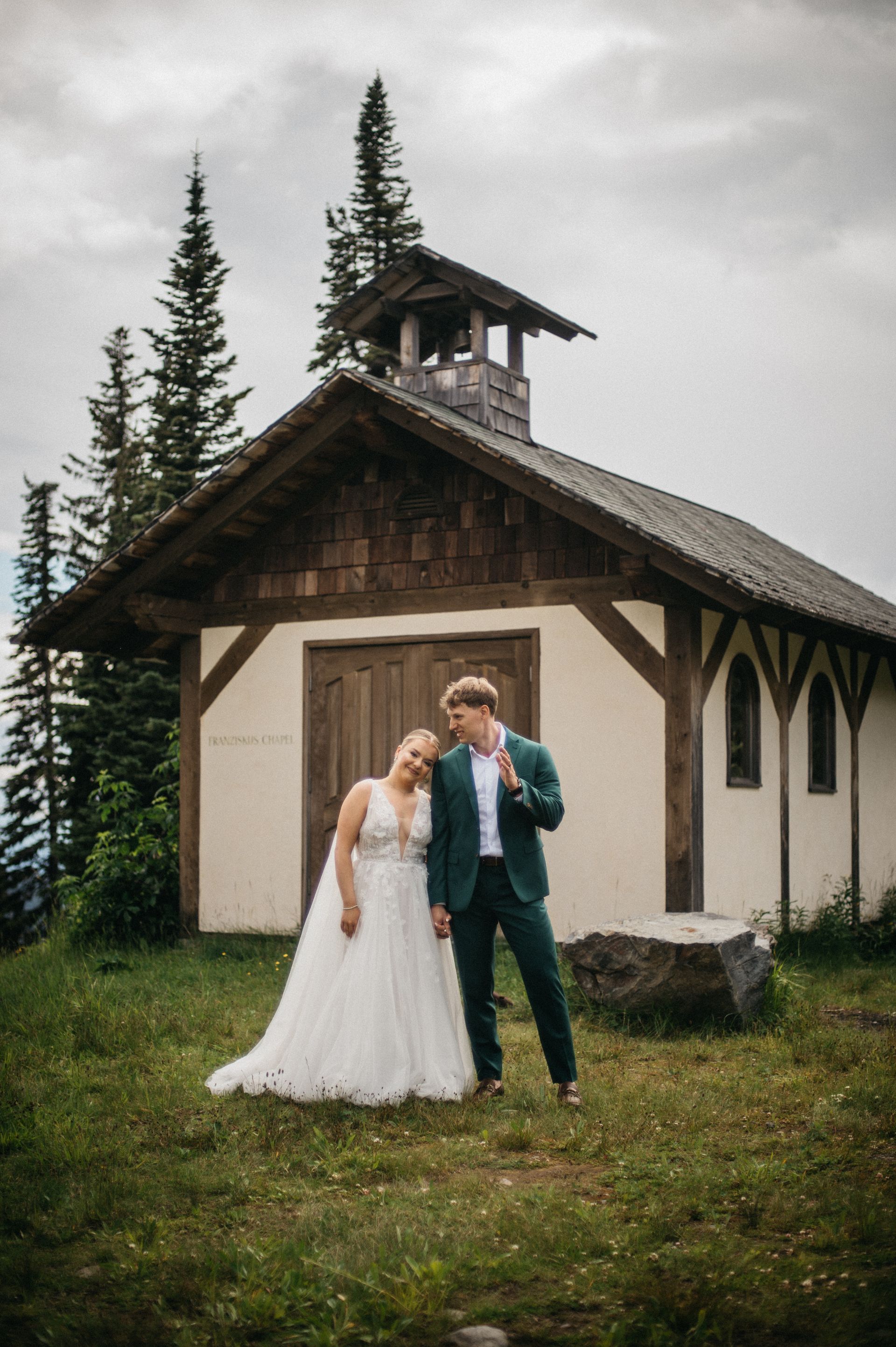 Couple in wedding attire posing near a small chapel in a mountain setting.