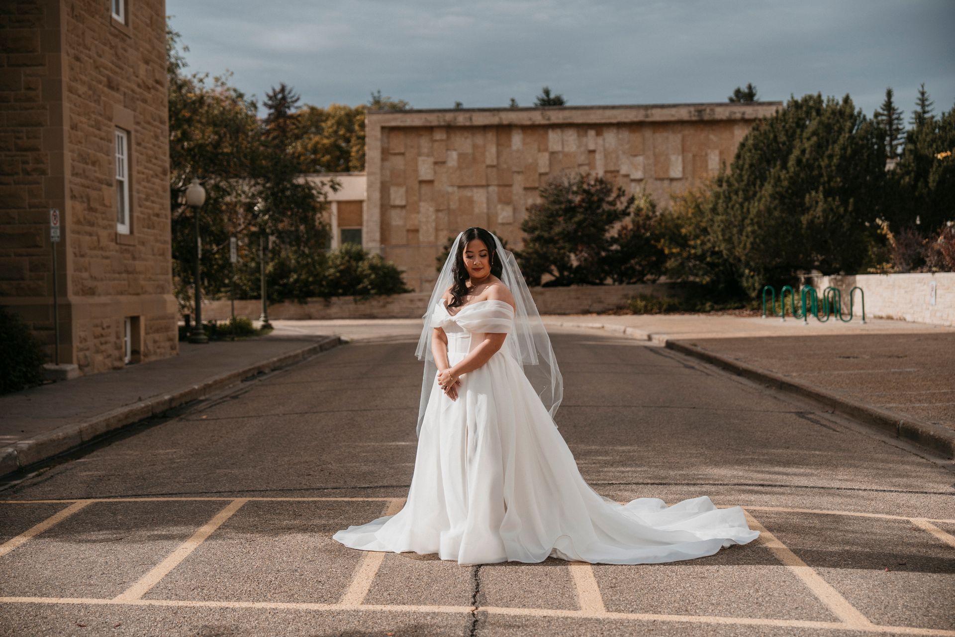 Bride in white dress with veil stands on a street with buildings and trees in the background.