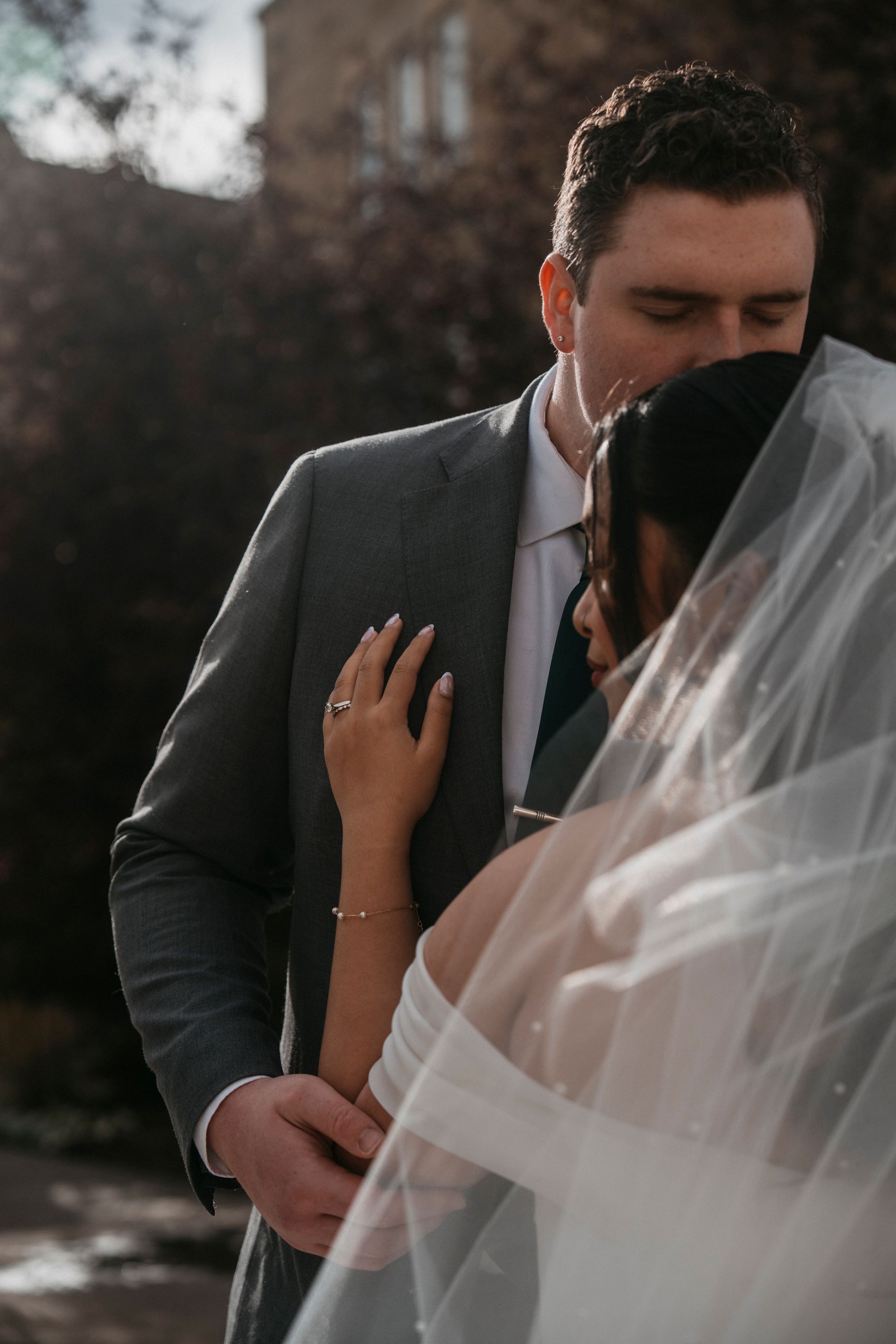 Couple embraces outdoors; man in gray suit, woman in white wedding dress with veil, sunlight.