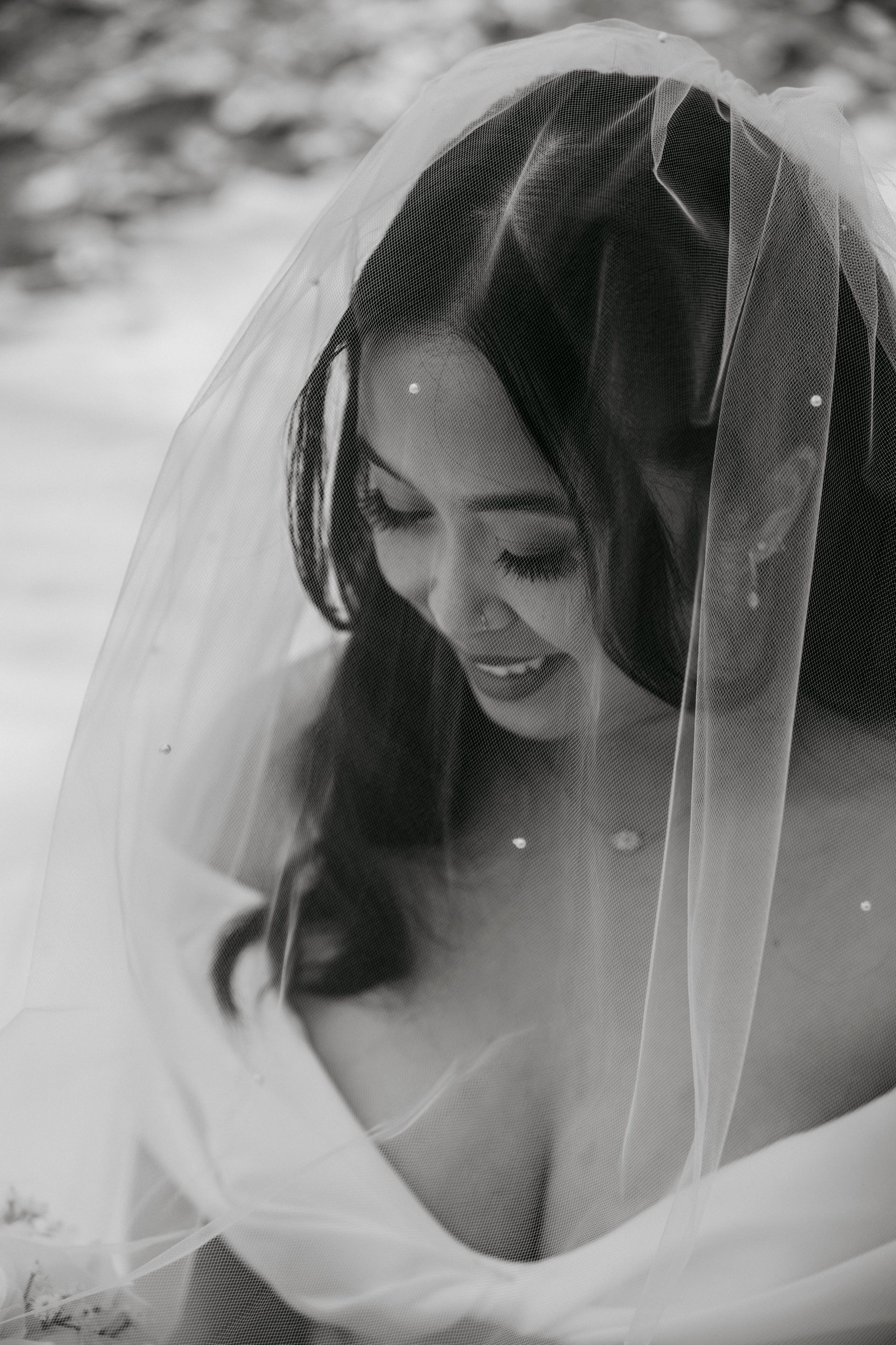 Woman in a veil, looking down. Black and white photograph, outdoor setting, possibly a wedding.