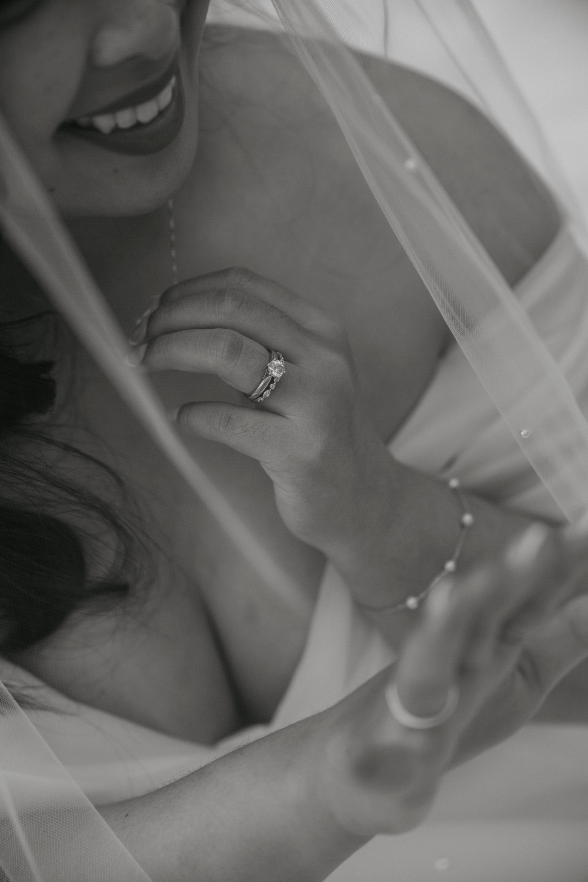 Bride adjusting necklace, ring on finger visible, veil draped over shoulder. Black and white.