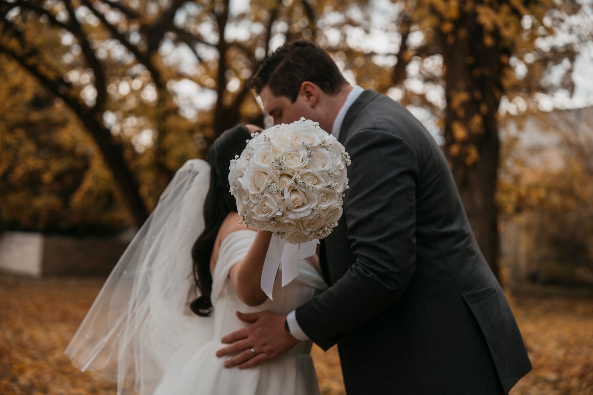 Bride and groom kissing, holding bouquet, outdoors with autumn foliage.