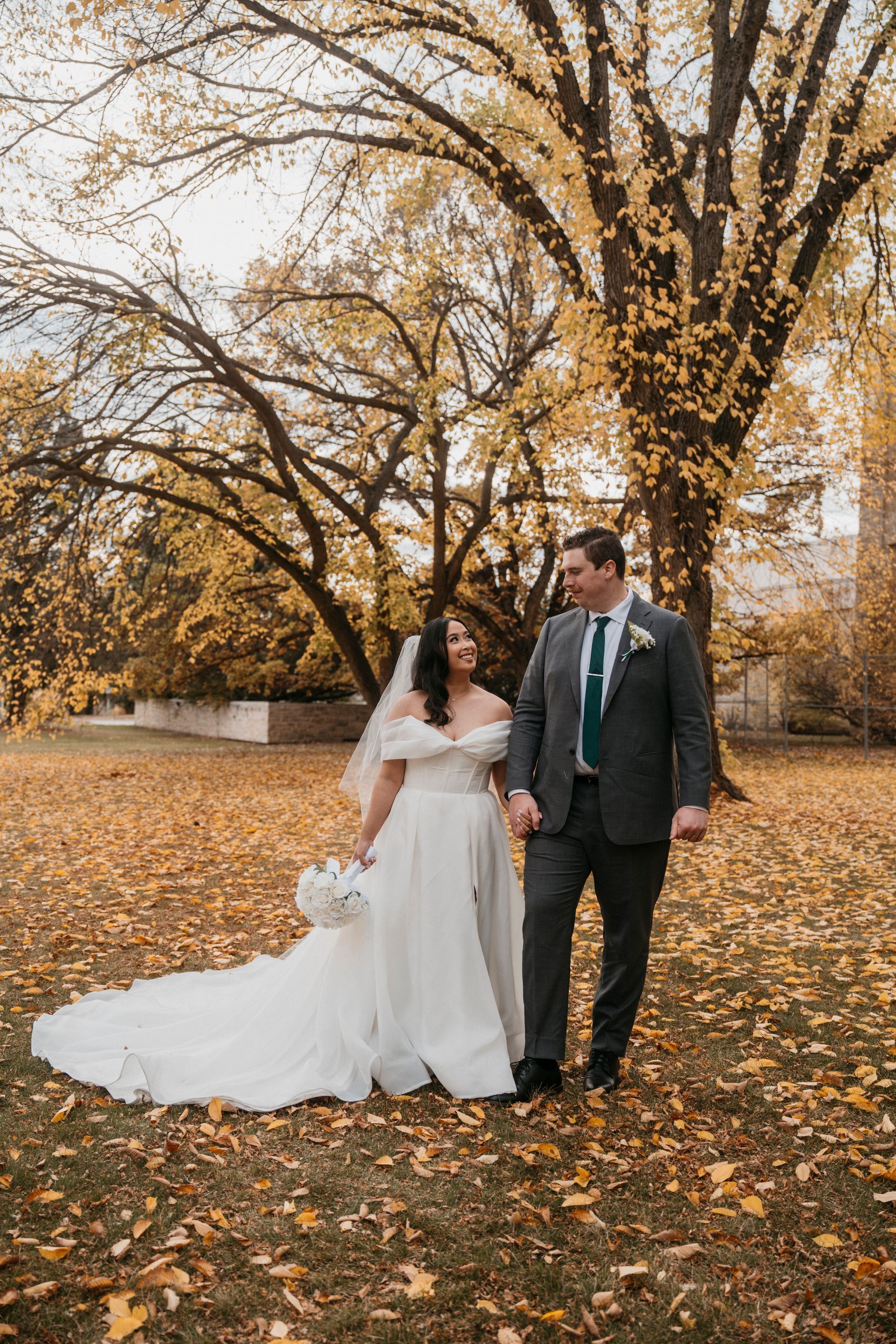 Newlyweds walking hand-in-hand in a park with golden fall foliage. Bride in a white dress and veil, groom in a gray suit.