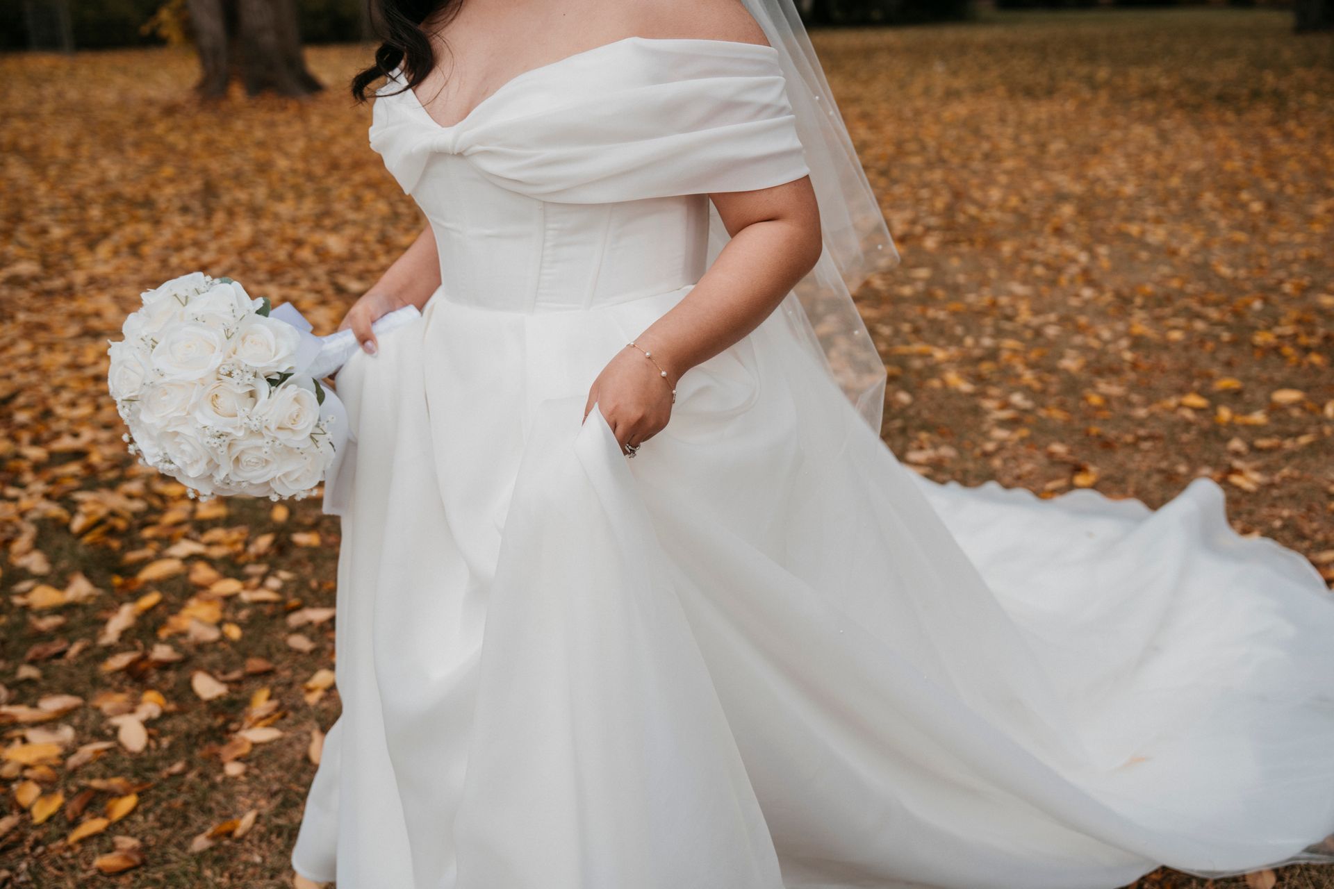 Bride in white off-the-shoulder gown, holding a bouquet. Outdoors with fall leaves.