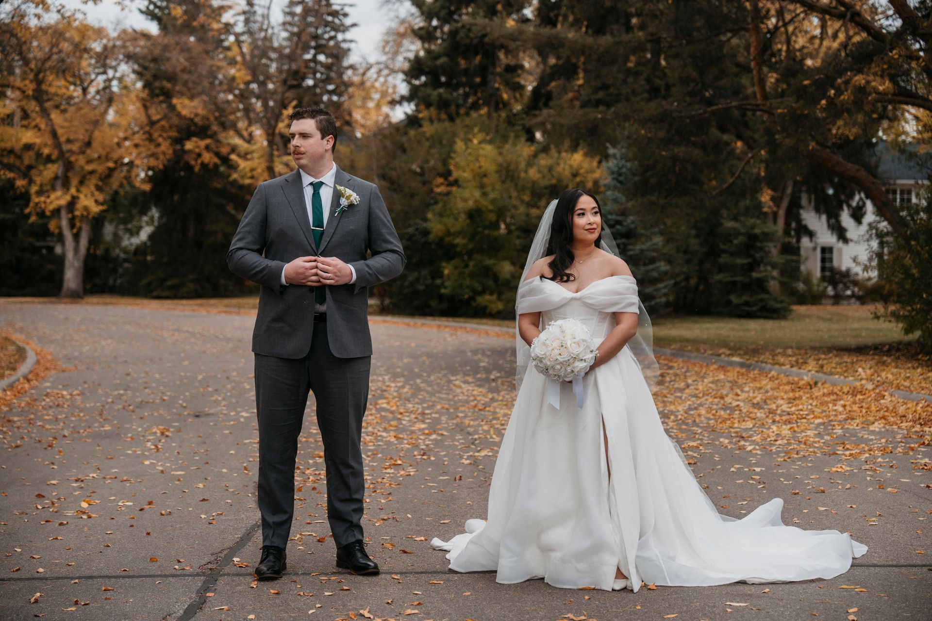 Bride and groom pose on a road lined with fall foliage. The groom wears a gray suit, and the bride is in a white dress with a bouquet.