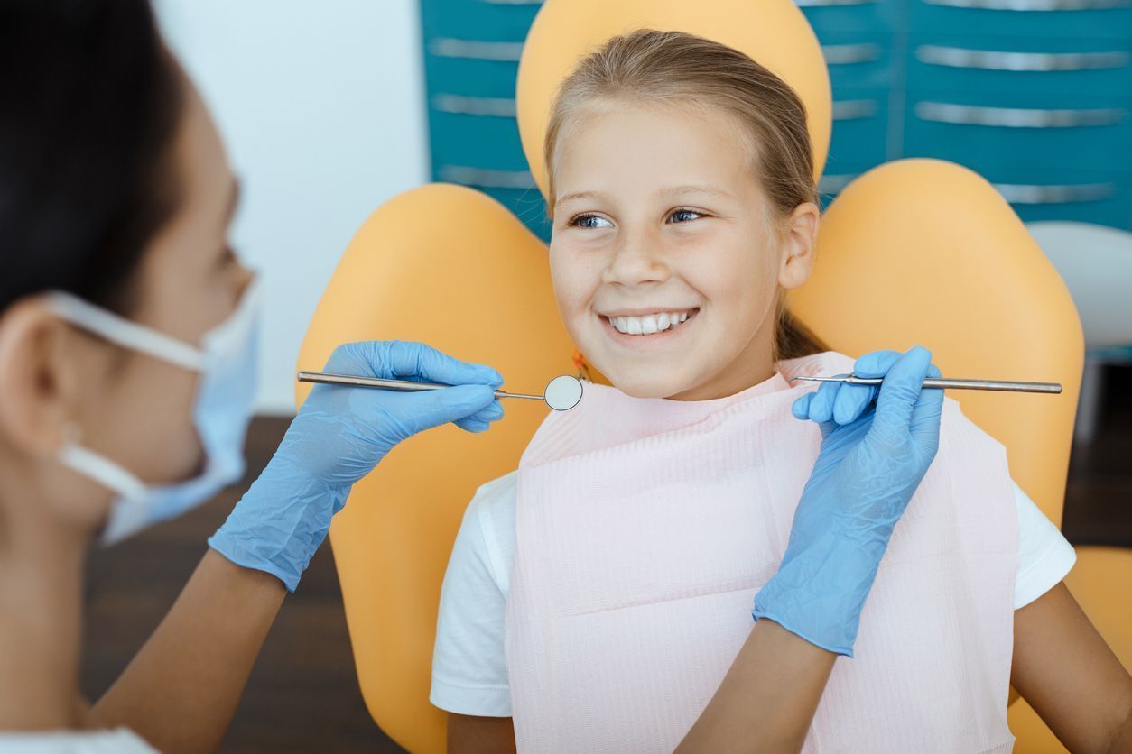A dental professional in blue gloves holds a mirror while examining the teeth of a smiling patient in a yellow chair.