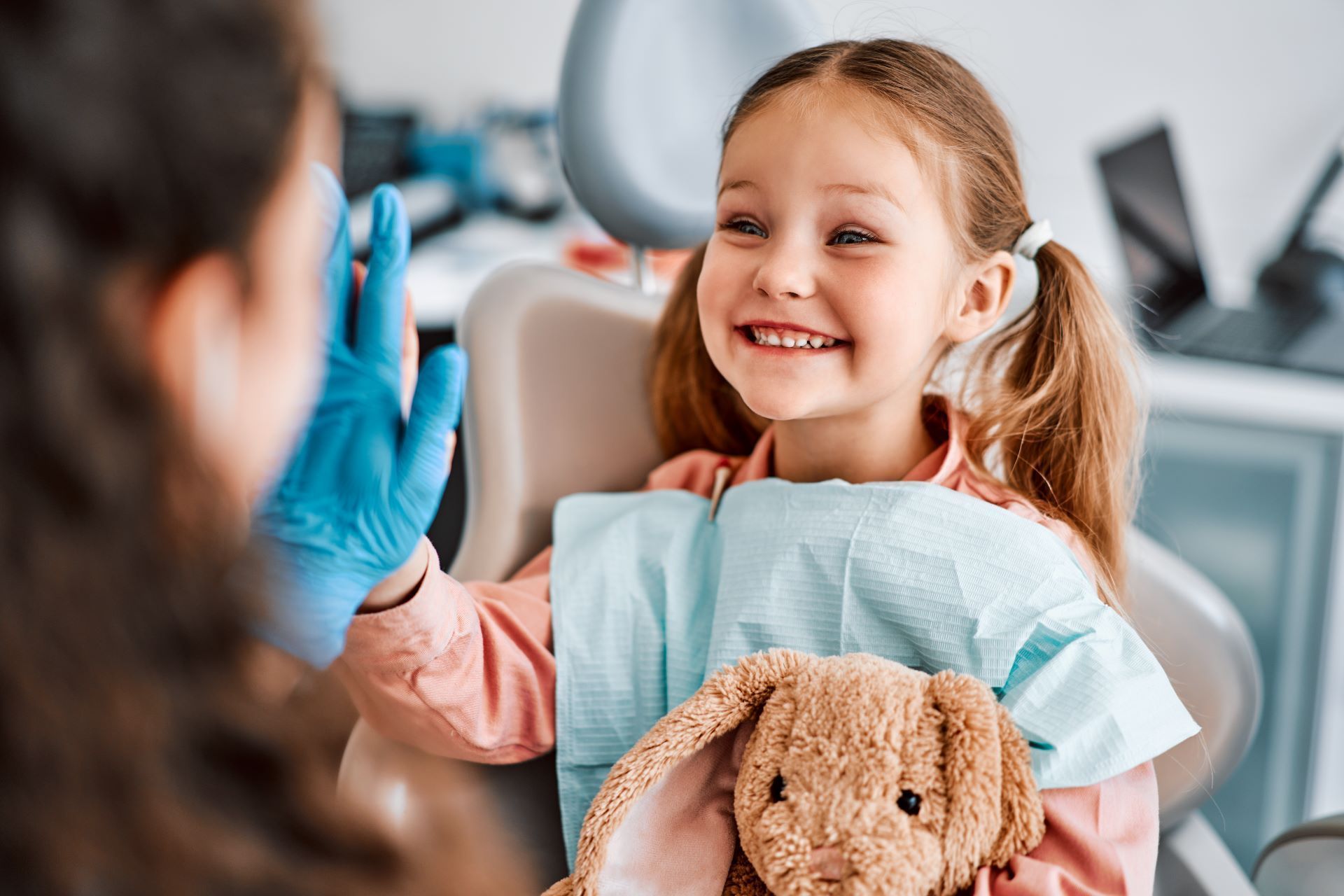 A person wearing blue medical gloves high-fives a smiling patient holding a stuffed toy in a dental chair.
